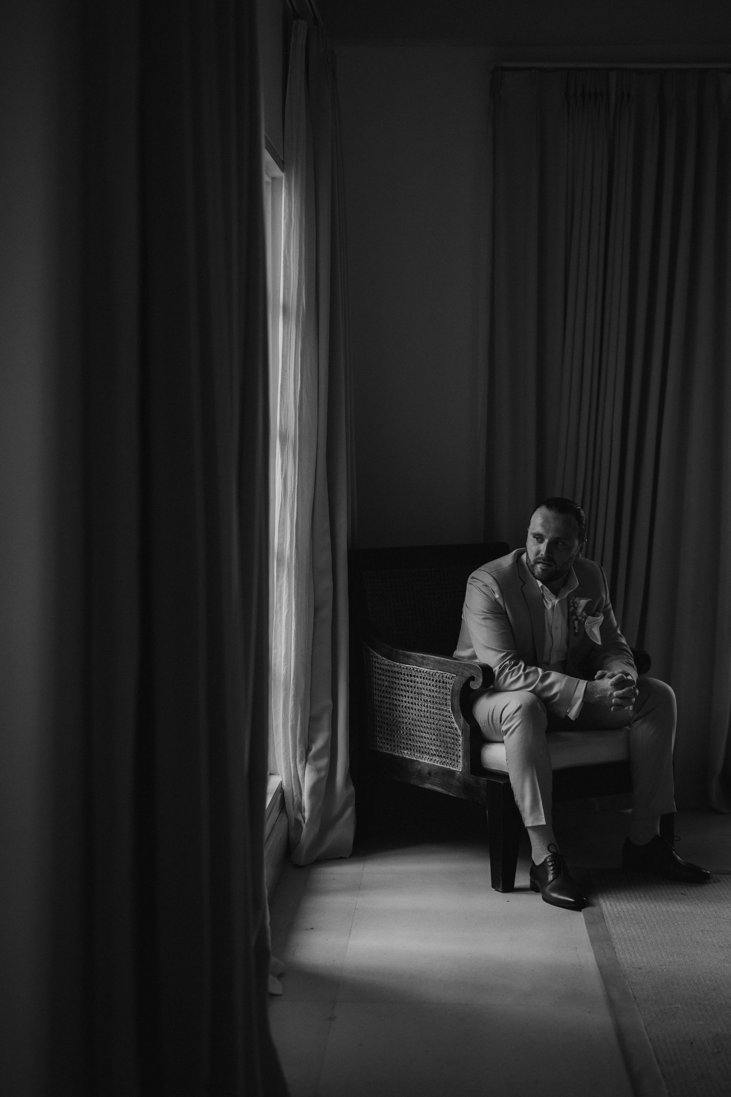 Black and white photo of a groom in a suit sitting alone in a dimly lit room by curtains.