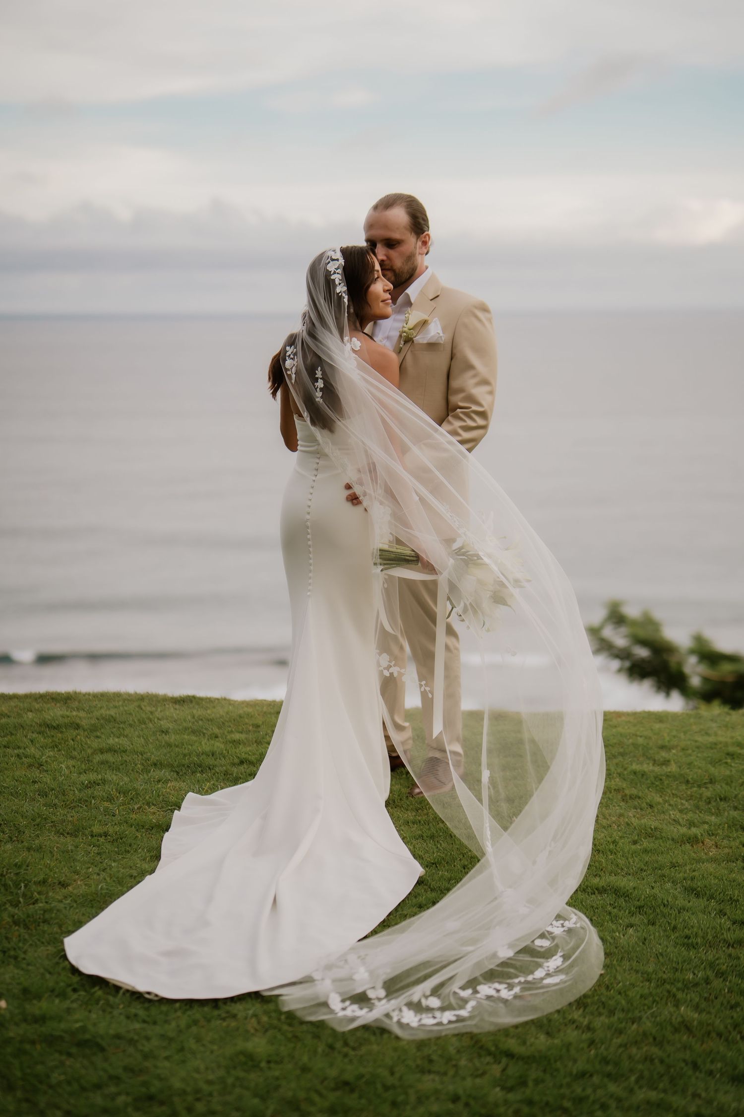 Bride and groom kissing with flowing lace veil billowing in ocean breeze.