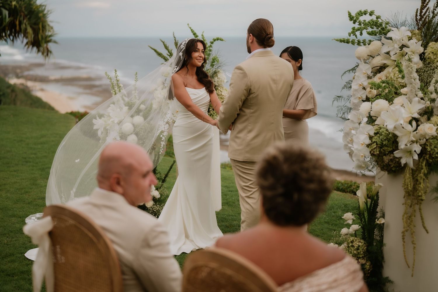 Bride and groom exchange vows at outdoor oceanfront ceremony surrounded by lush white floral arrangements.