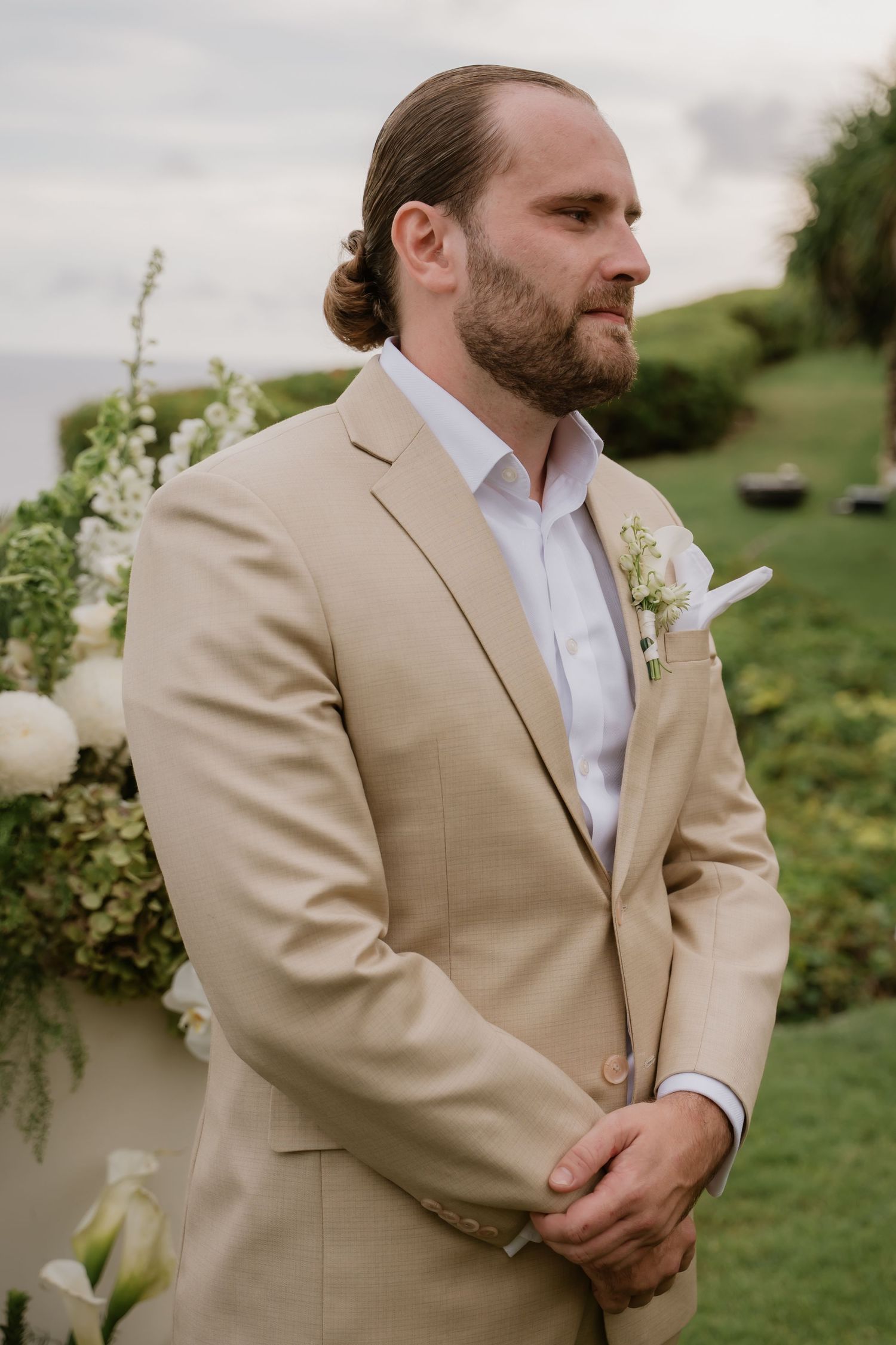 Groom in beige suit with boutonniere stands at outdoor wedding ceremony overlooking the ocean.