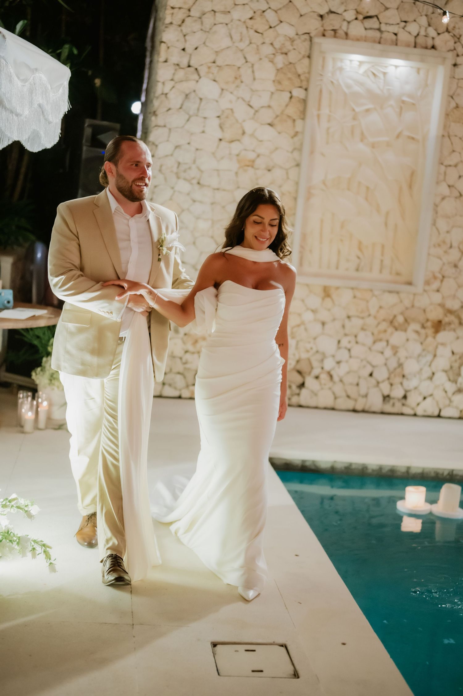 Bride and groom walking poolside at night in white gown and tan suit at wedding reception.