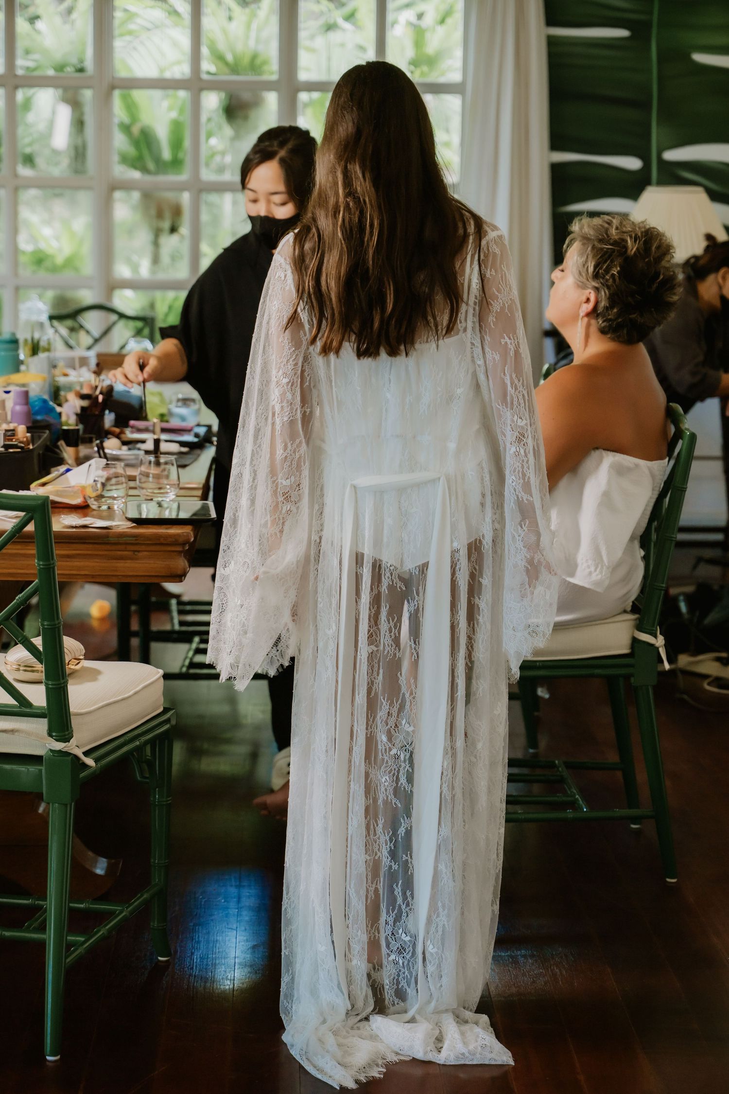 Bride in sheer lace robe getting ready with hair and makeup artists in a tropical venue.