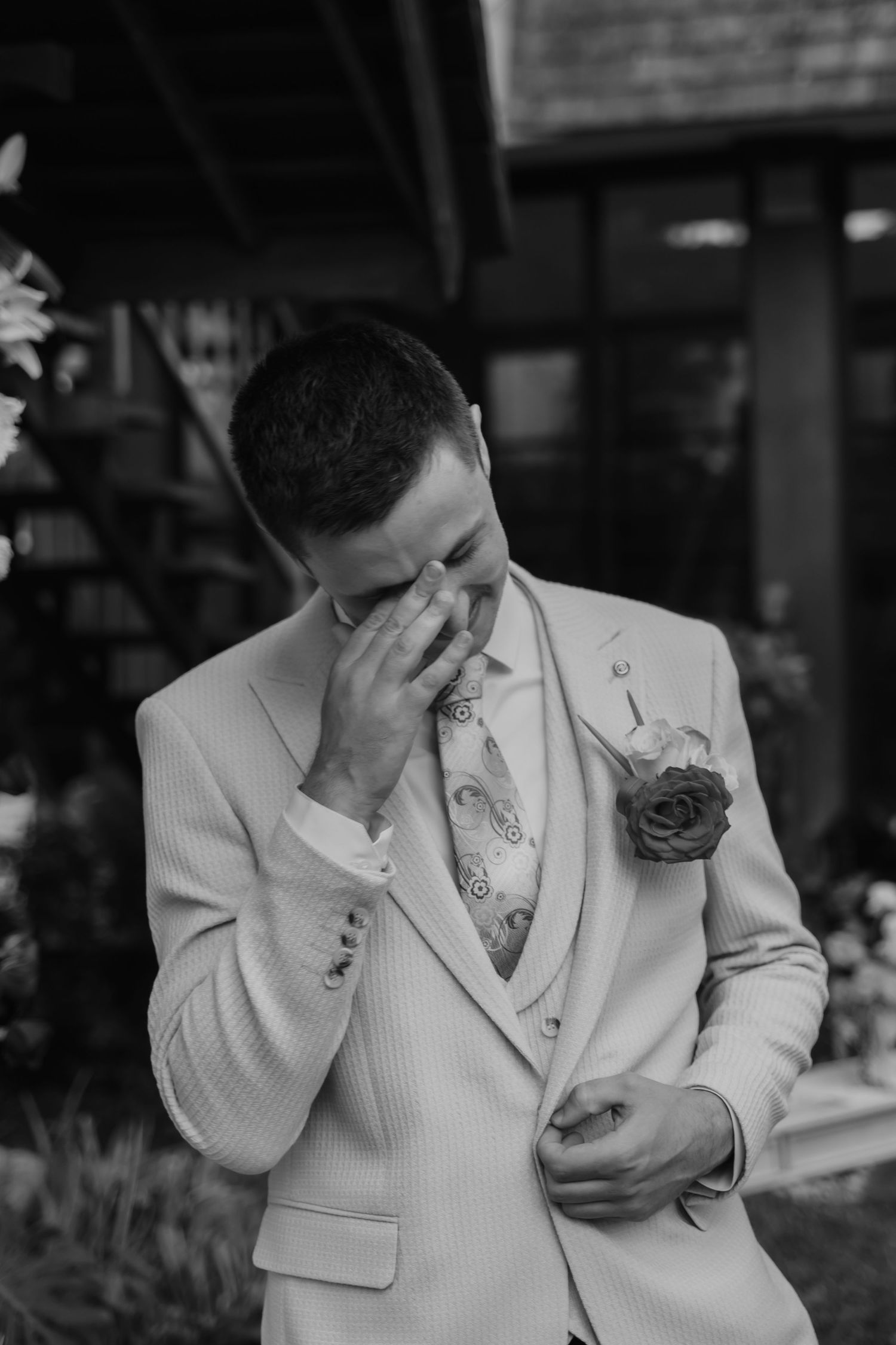 Emotional groom in white suit wiping tears with boutonniere on lapel during wedding ceremony outdoors.