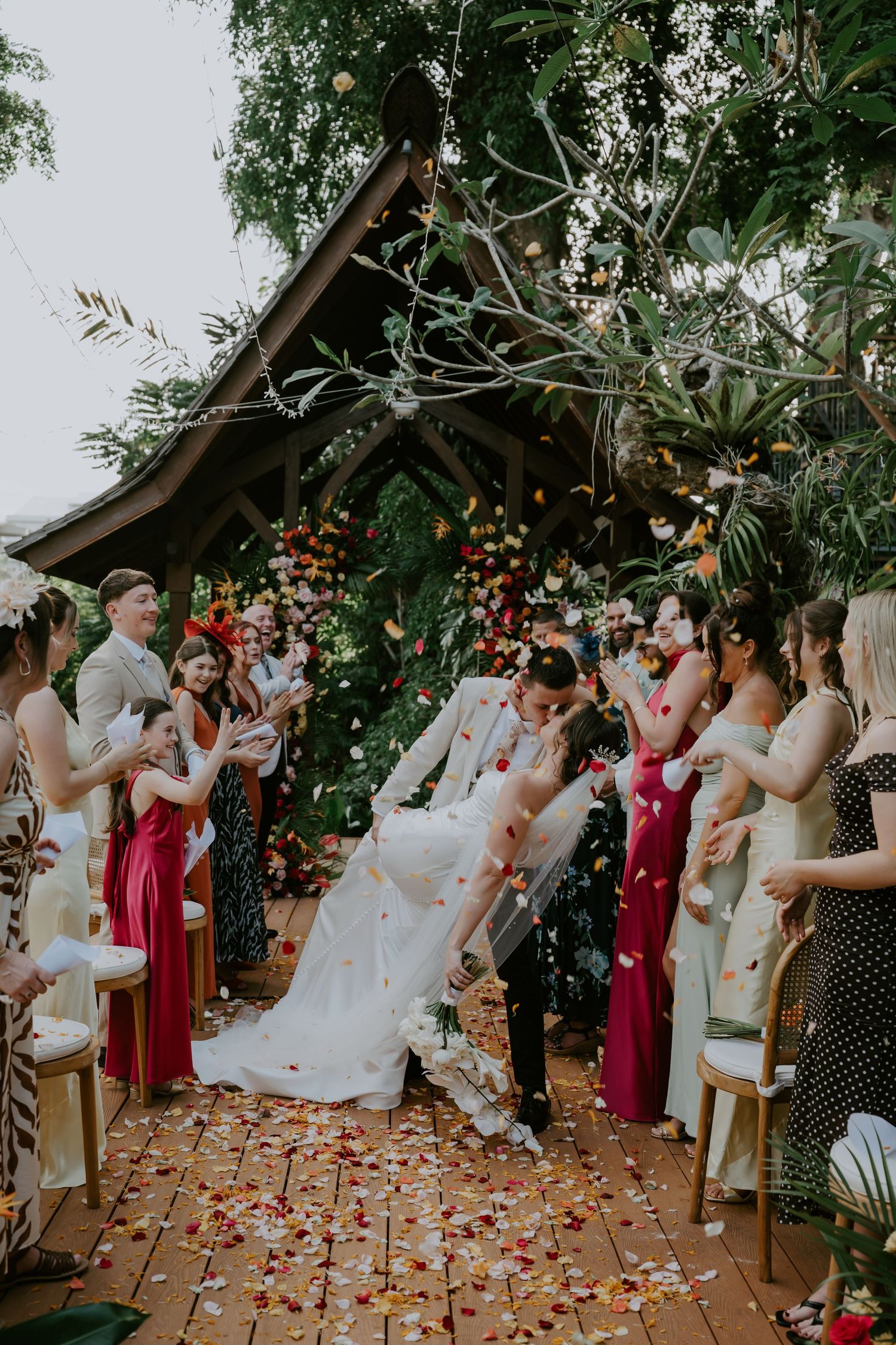 Couple shares a kiss as guests toss colorful flower petals at a lush tropical wedding ceremony.