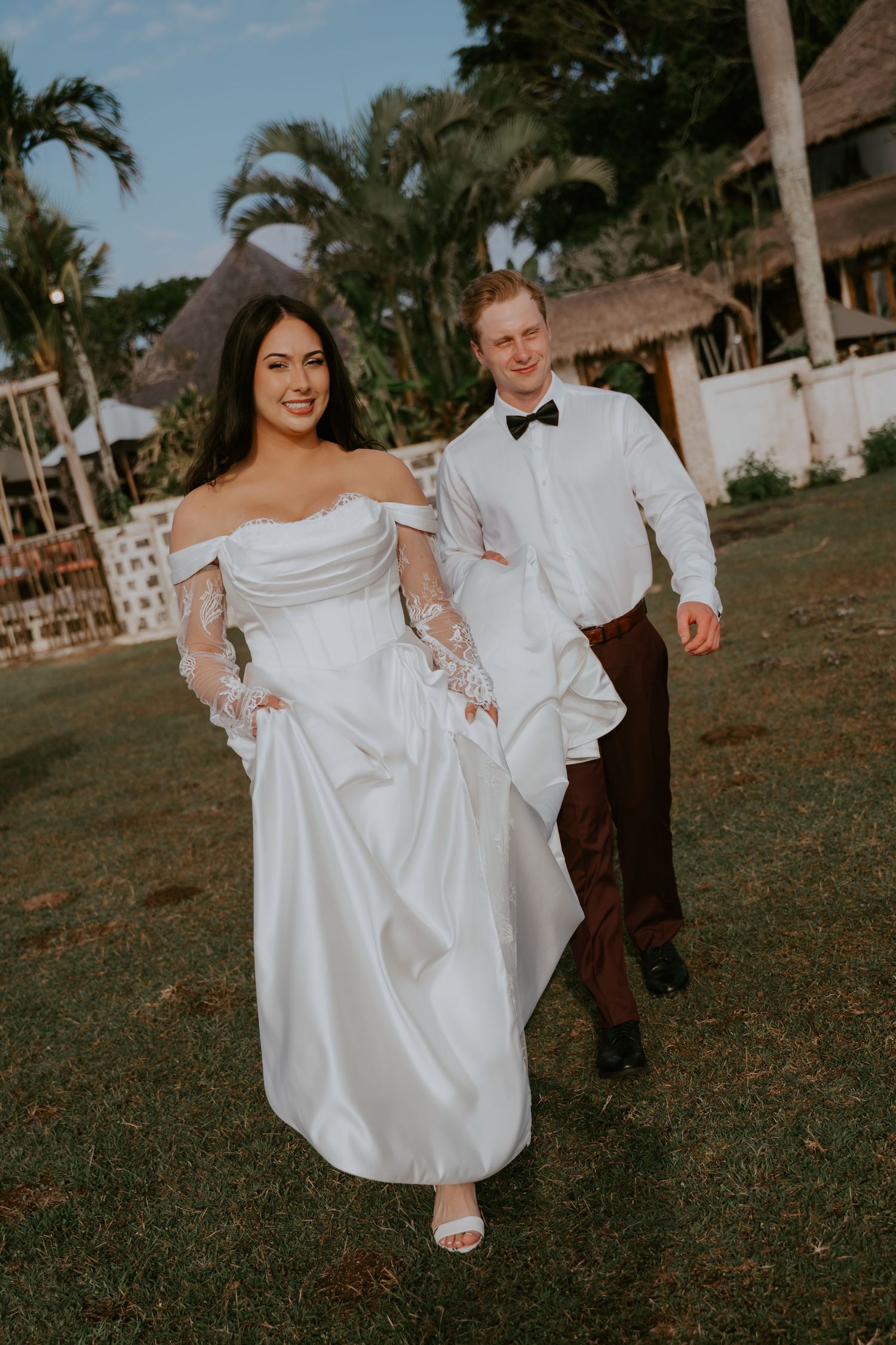 Smiling bride in off-shoulder lace wedding gown walks with groom in white shirt and bow tie on green lawn.