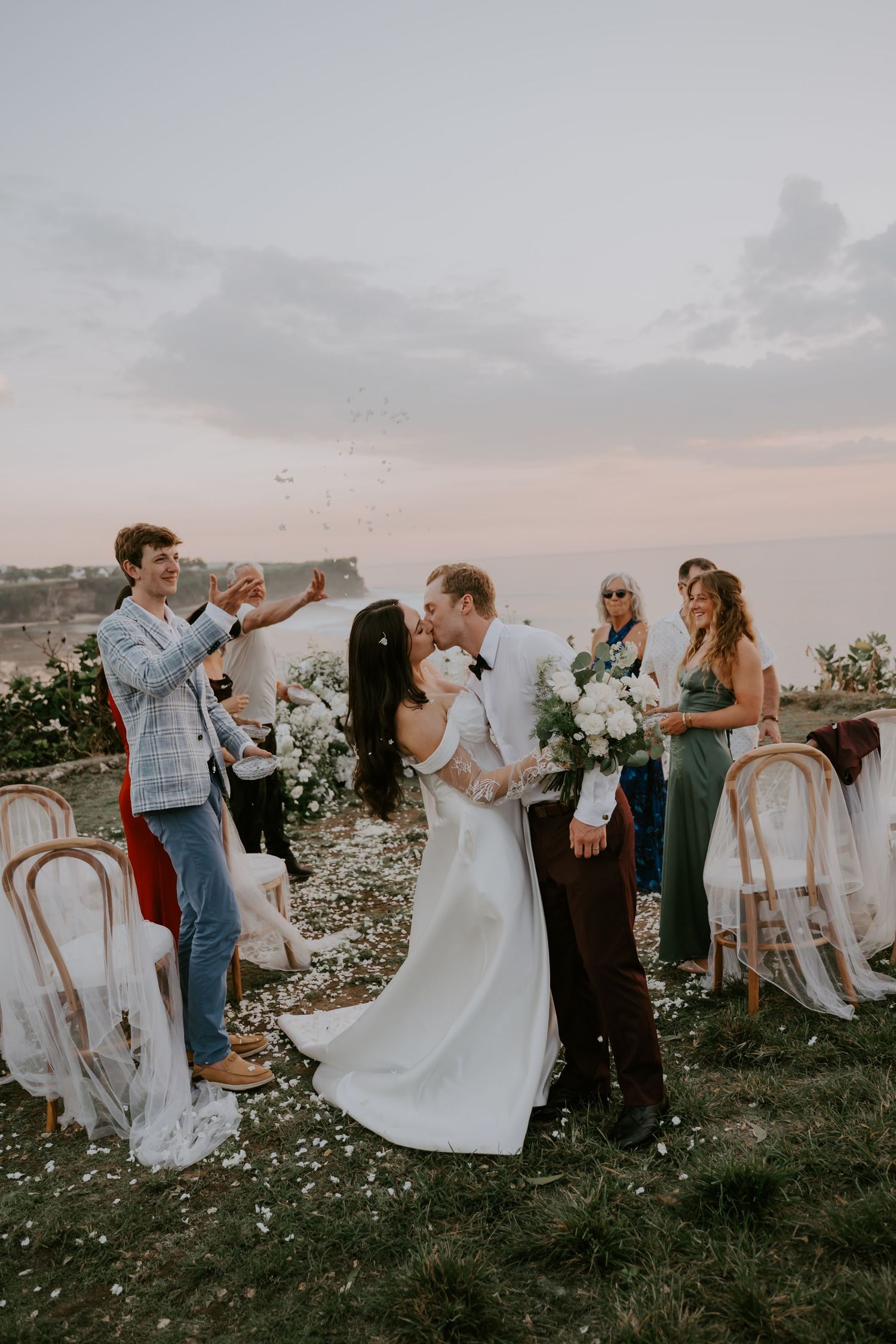 Bride and groom share first kiss as guests celebrate with confetti at outdoor coastal wedding ceremony.