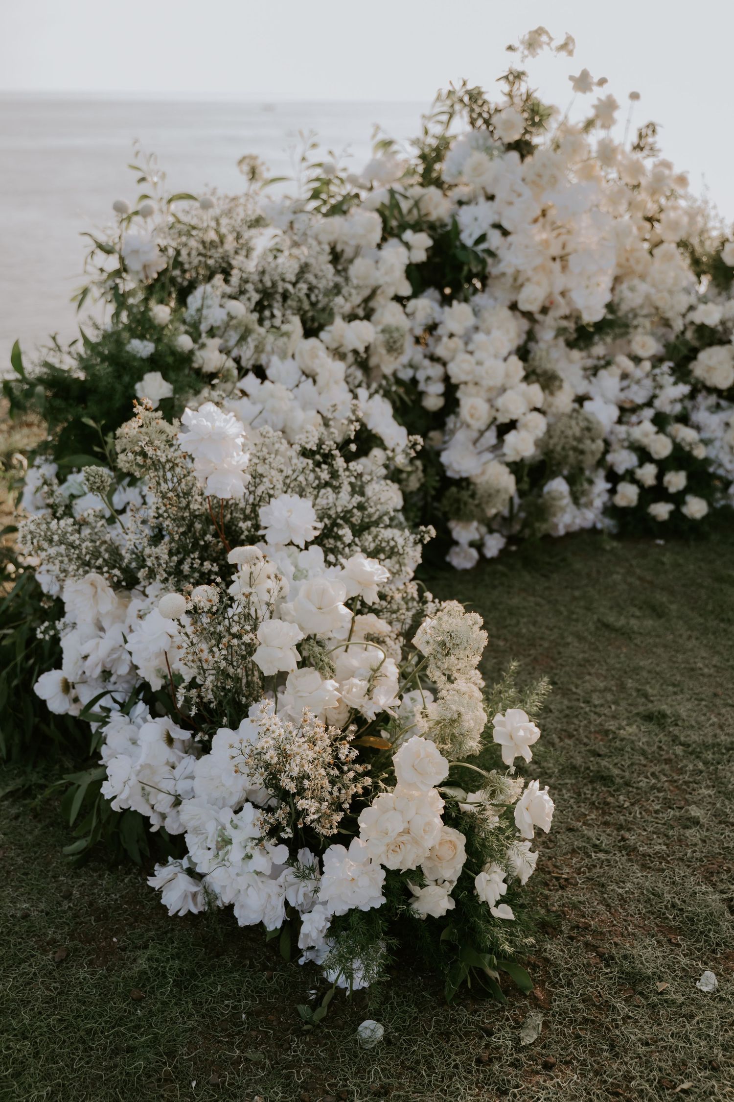 Lush white floral arrangements featuring roses and baby's breath cascading on grass at outdoor wedding.