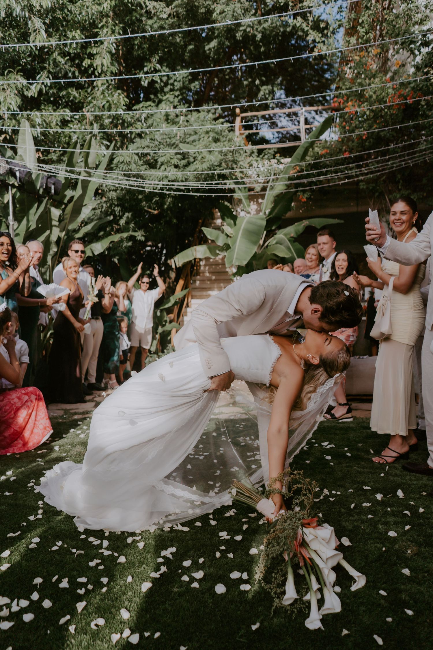 Groom dipping bride for romantic kiss on rose petal aisle surrounded by cheering wedding guests outdoors.