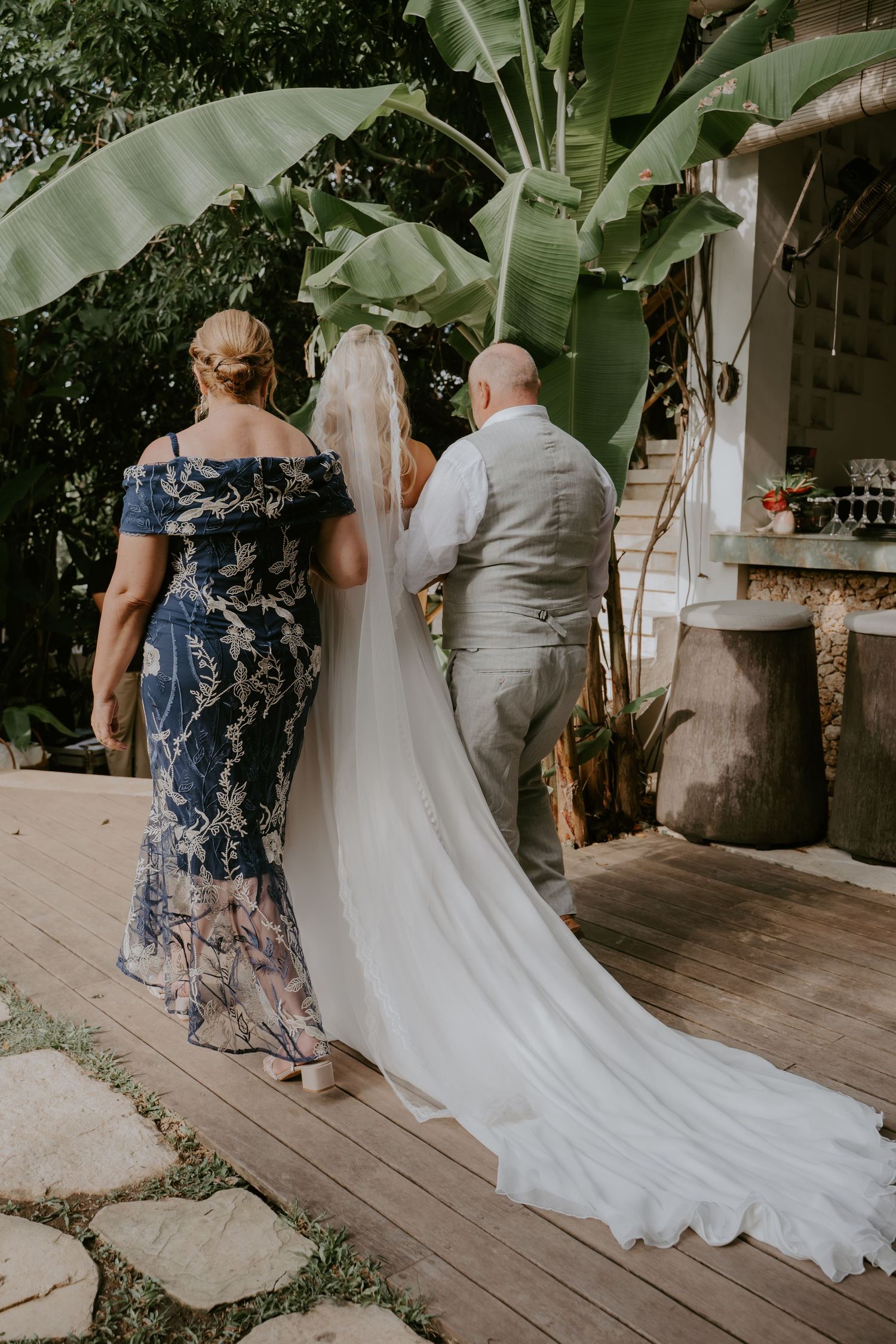 Bride in white gown and veil walks with parents on wooden deck surrounded by tropical greenery.