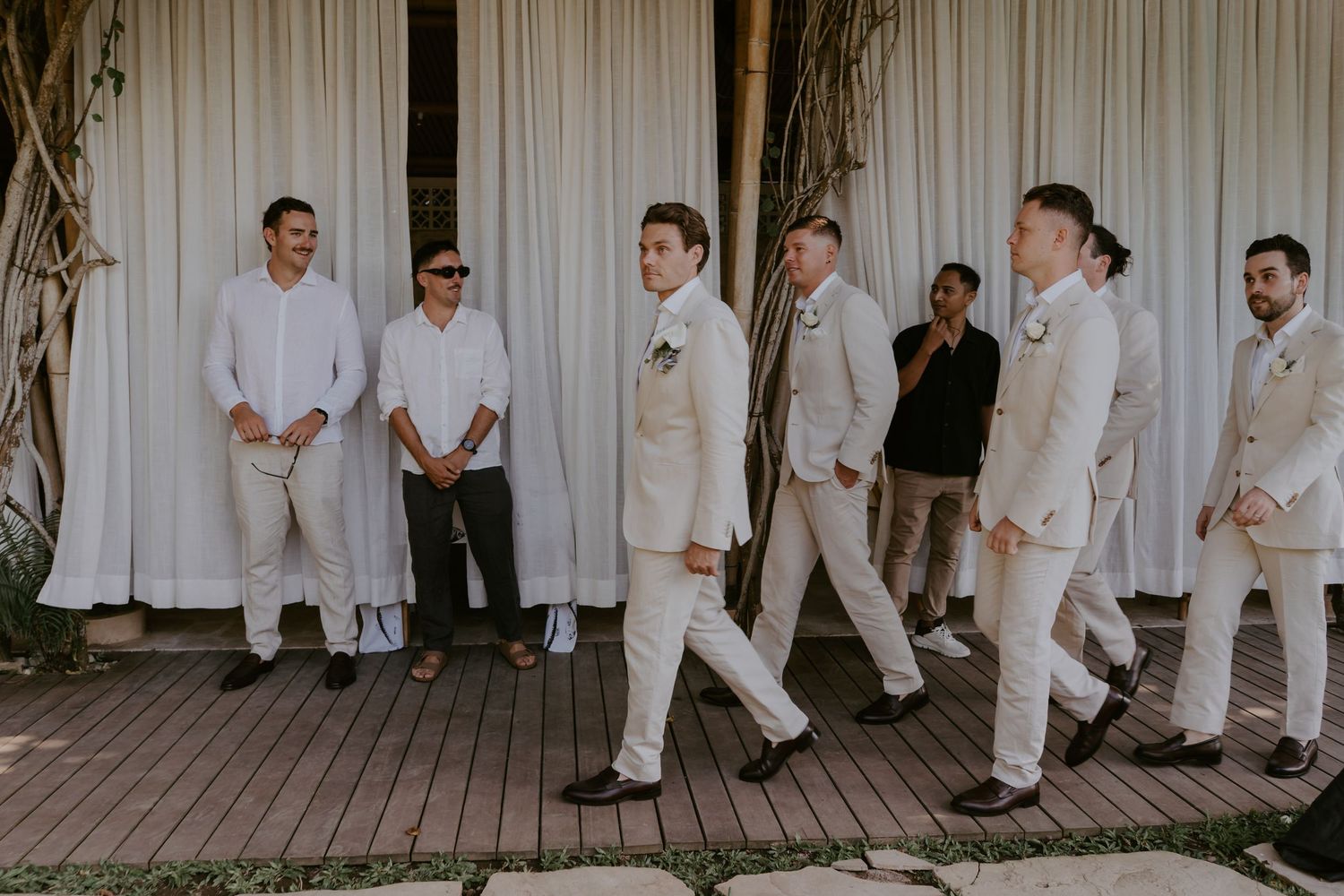 Groom and groomsmen in beige linen suits walking confidently along a wooden deck before the ceremony.