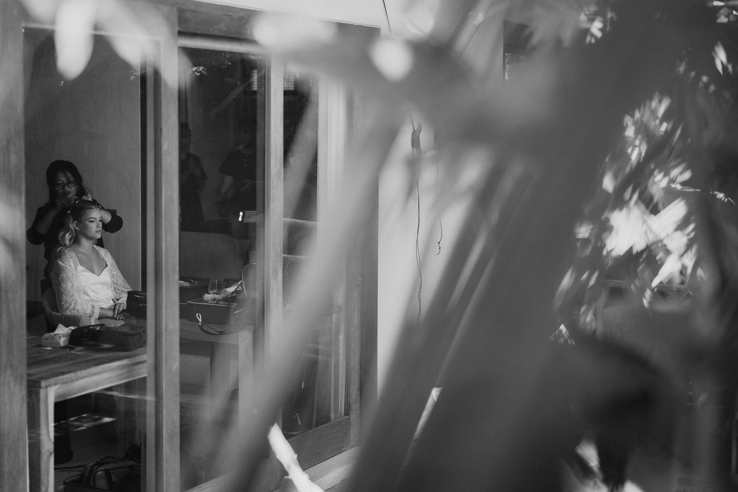 Black and white photo of a bride getting ready, viewed through a window with foliage in the foreground.