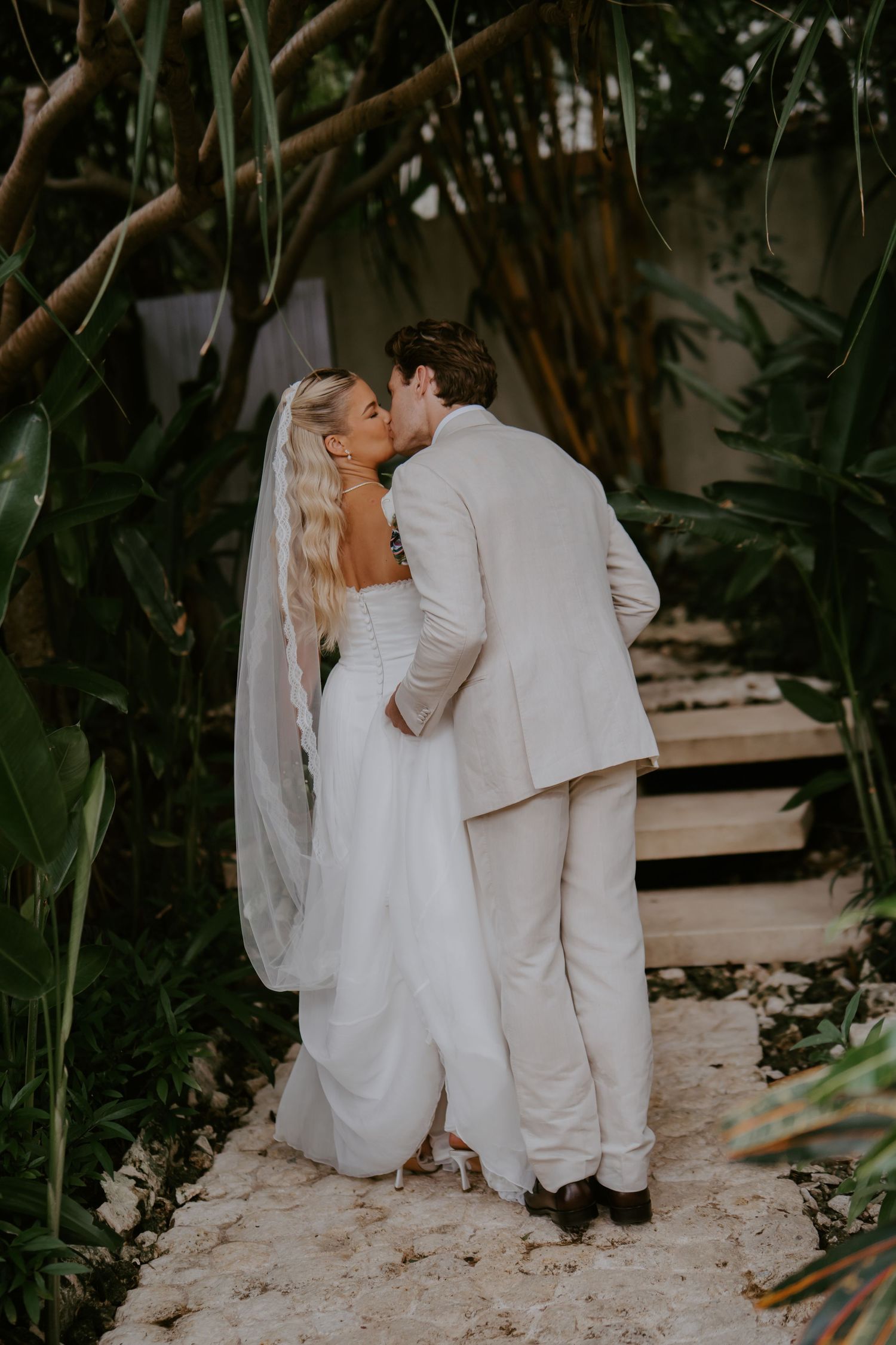 Bride and groom share a kiss on sandy garden path surrounded by lush tropical vegetation.