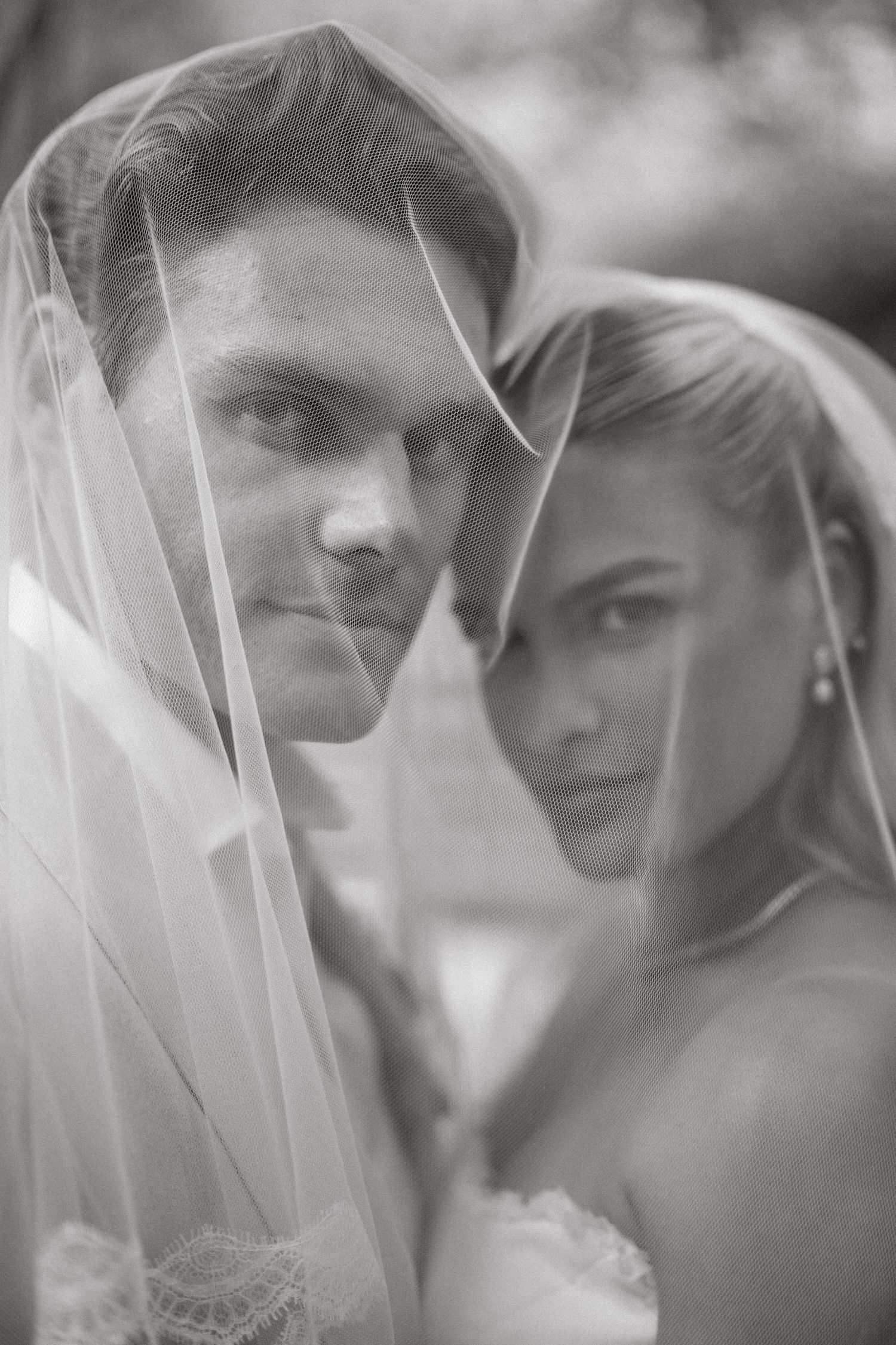 Black and white wedding portrait of bride and groom sharing a veil, gazing at camera.