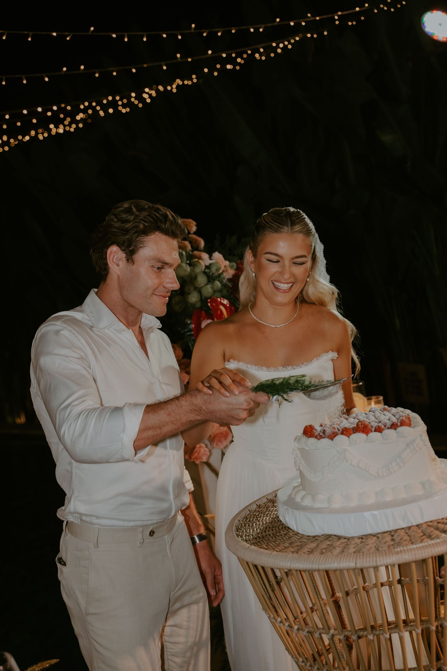 Smiling bride and groom cut their white wedding cake decorated with strawberries under romantic string lights at night.