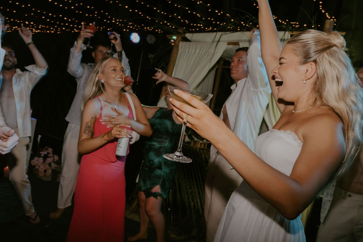 Bride laughs holding champagne glass while dancing with guests at wedding reception.