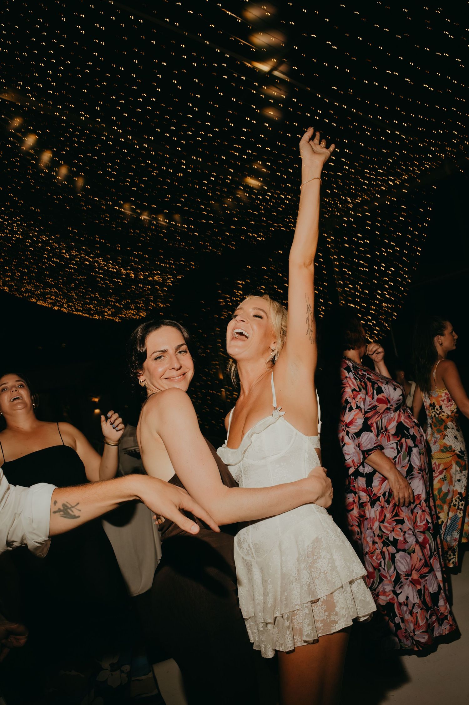 Bride laughing and dancing with arm raised at a glittering fairy light wedding reception.