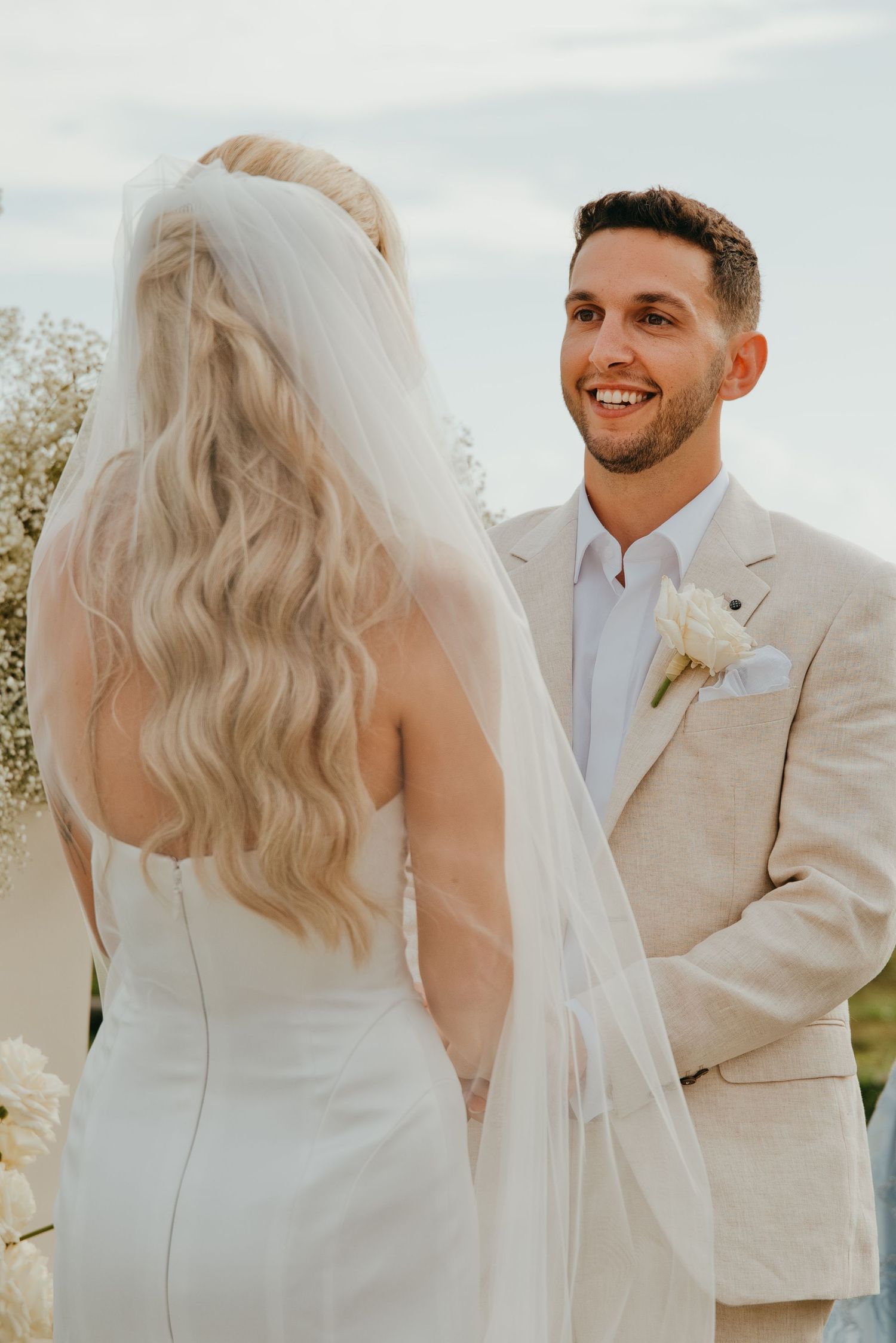 Smiling groom in beige suit with white boutonniere holding hands with bride during ceremony.