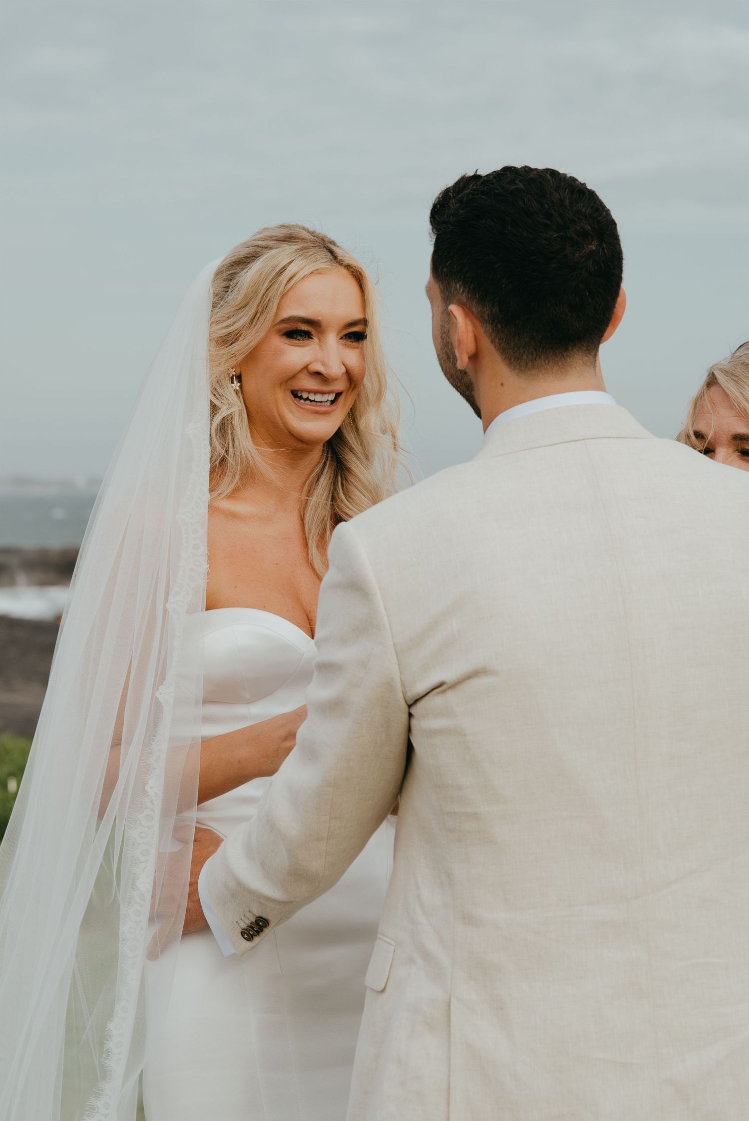 Radiant blonde bride smiling at groom during outdoor wedding ceremony with veil flowing behind her.