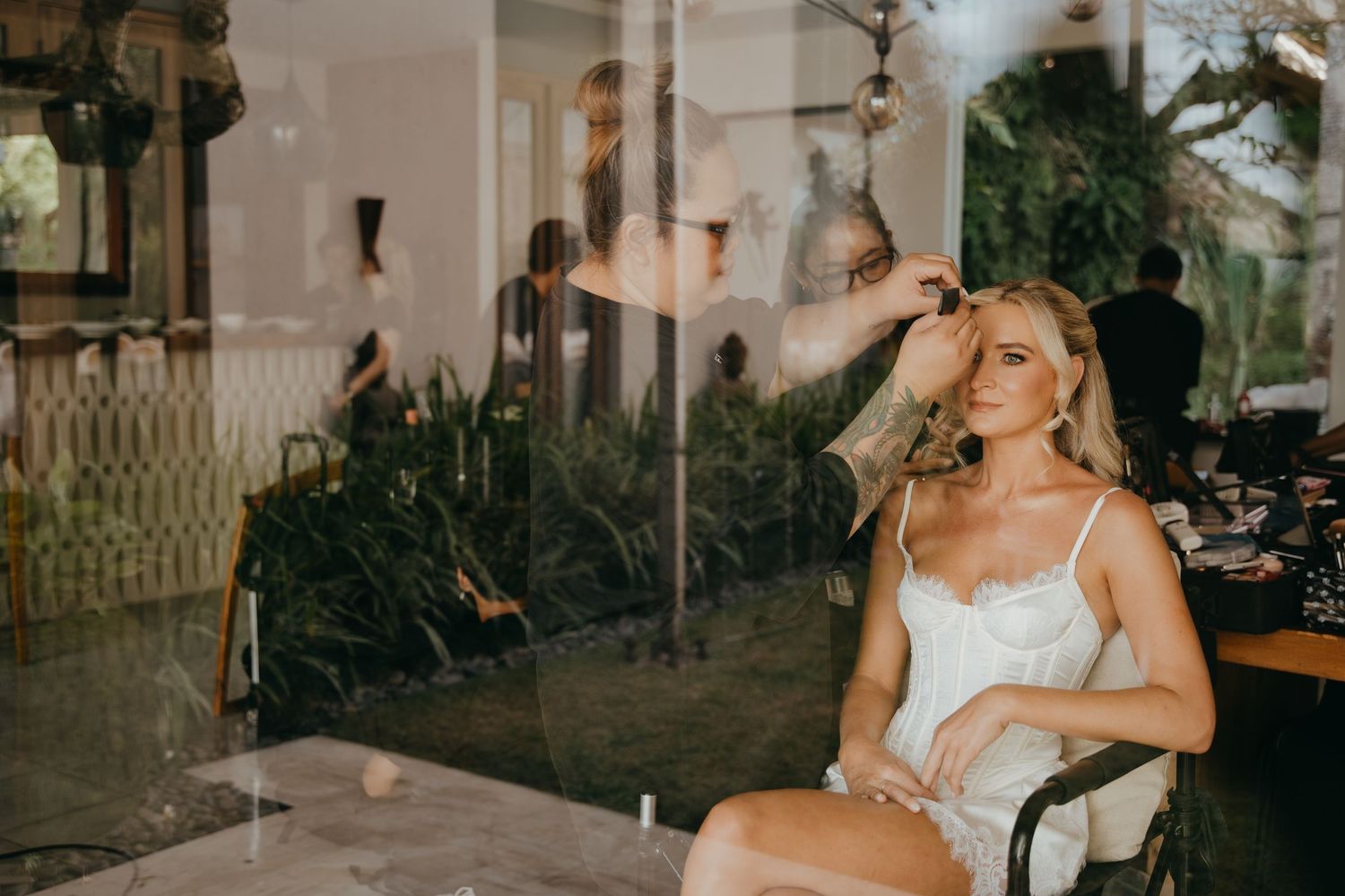 Woman in white dress sitting by glass window, smiling, with lush green garden backdrop in elegant setting.