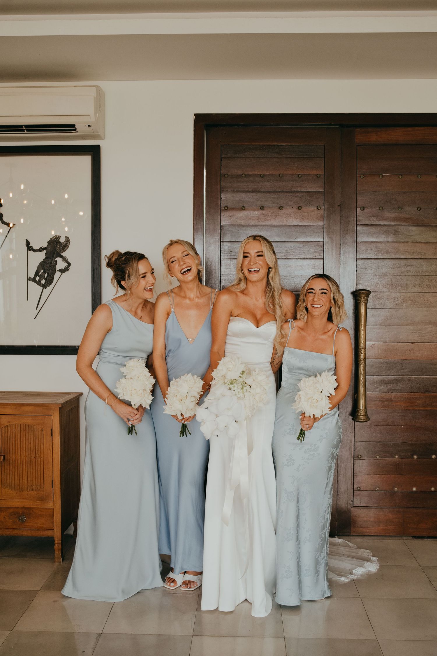 Bride in strapless white gown laughs with three bridesmaids in light blue dresses holding white bouquets.