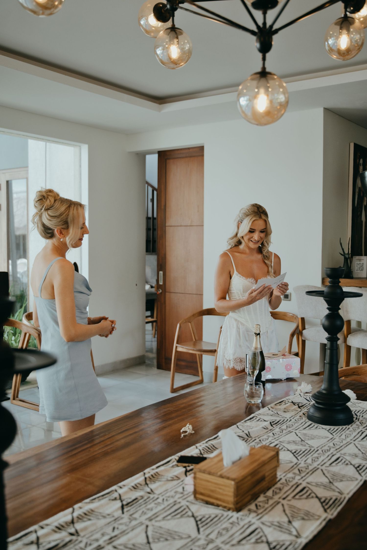 Bride in white lingerie reads a card while bridesmaid in grey dress looks on in a modern villa getting ready room.