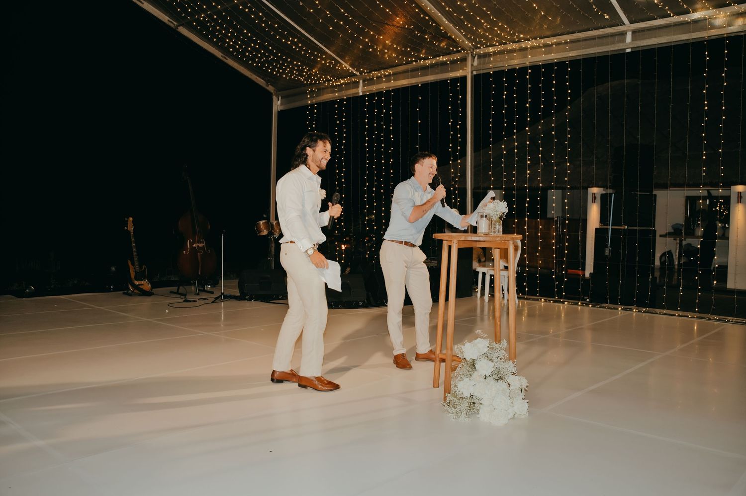 Two men giving speeches at a wedding reception on a white dance floor with fairy light backdrop.