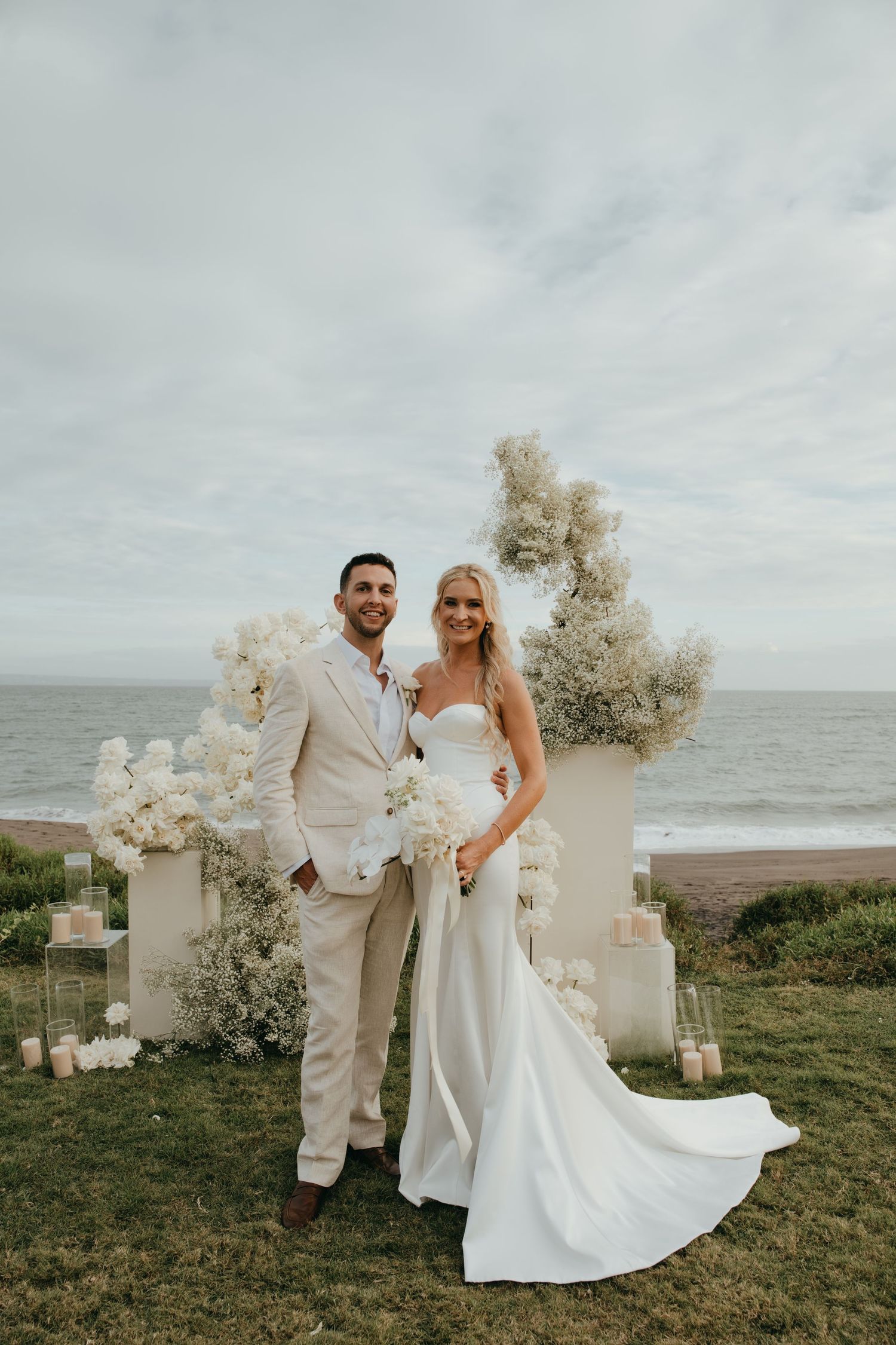 Bride and groom pose together before a white floral arch with ocean backdrop after ceremony.