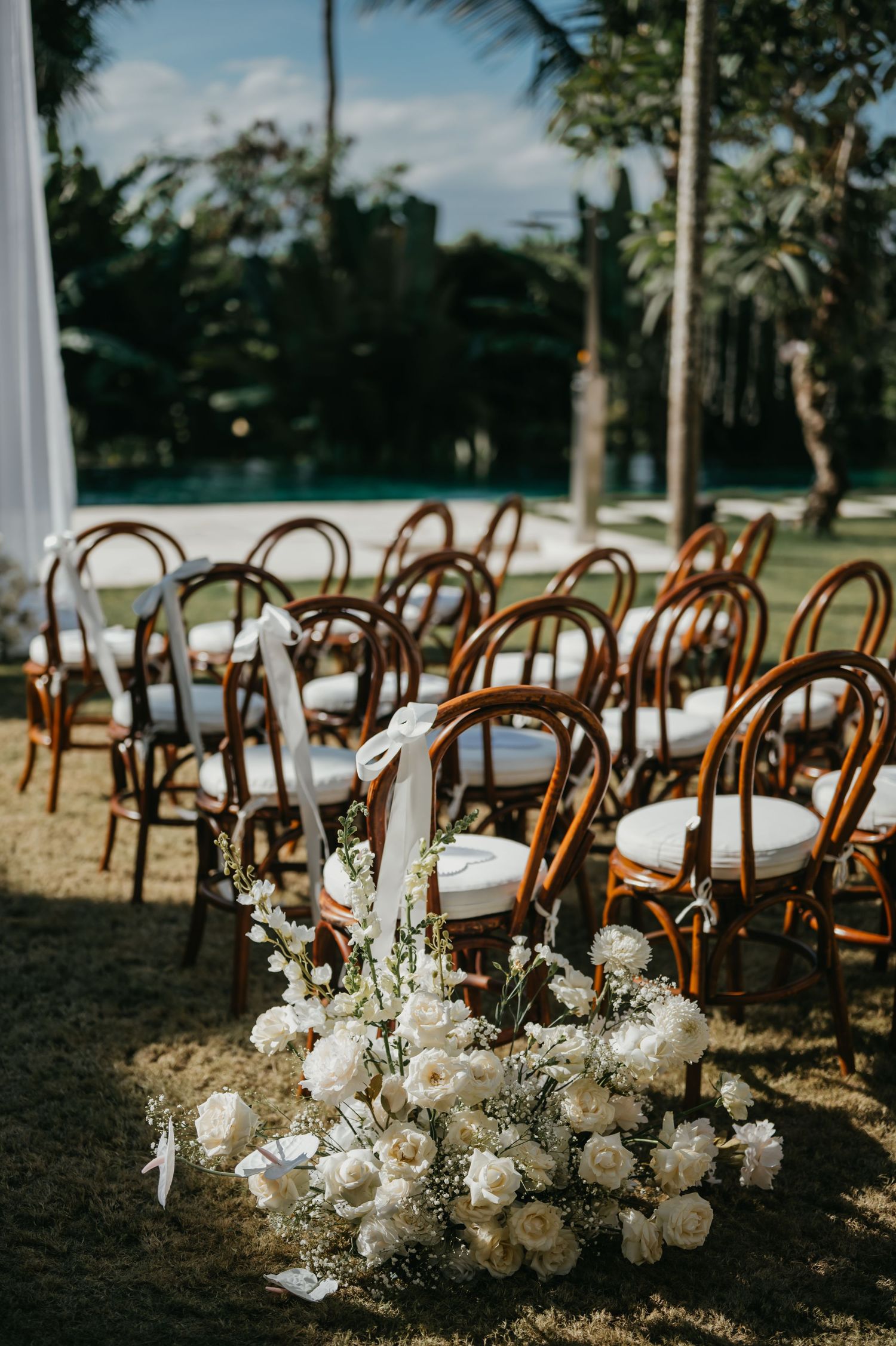 Outdoor wedding ceremony setup with bentwood chairs and white floral arrangements on a tropical garden lawn.