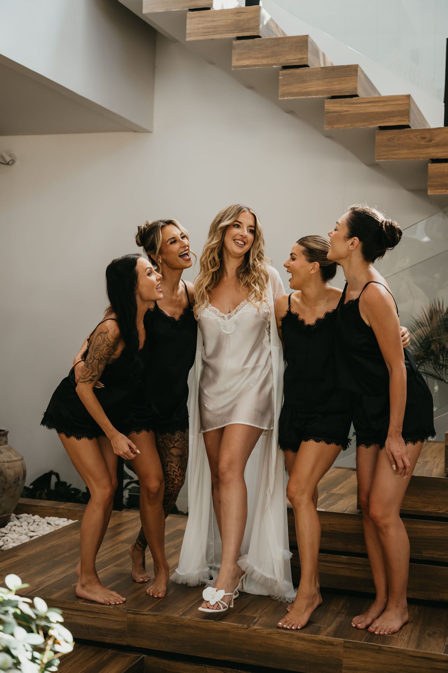 Bride in white silk robe with four bridesmaids in black lace sets on wooden staircase.