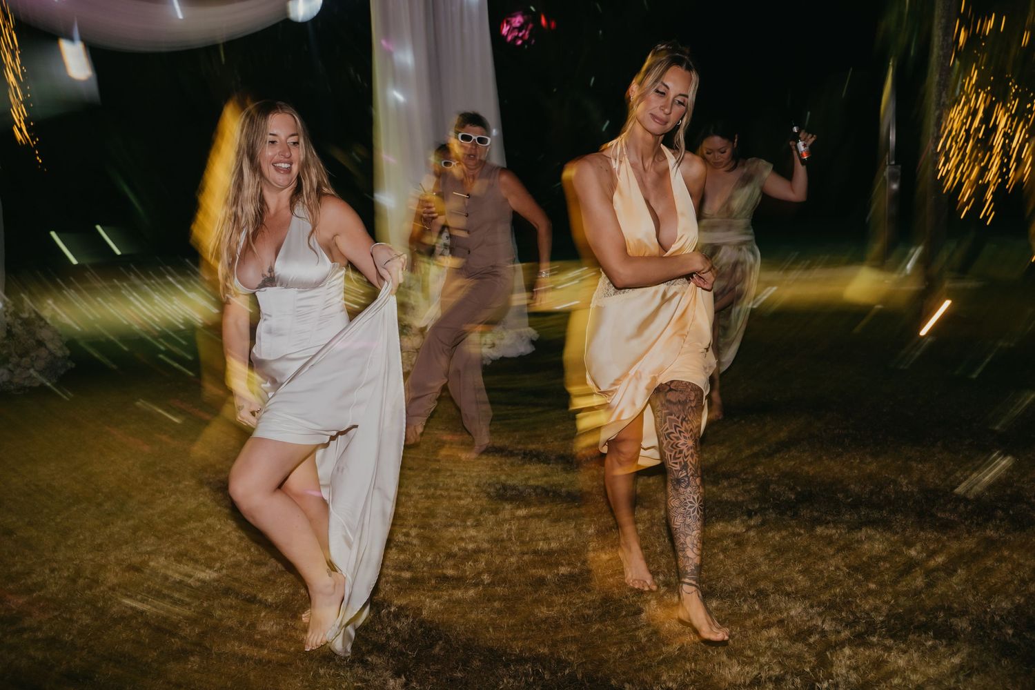 Guests in white dresses dance joyfully together at an outdoor tropical evening wedding reception.