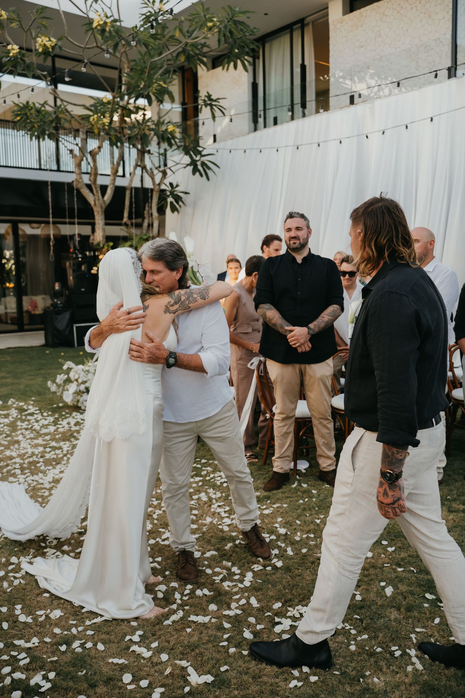 Bride embraces guest while groom watches during outdoor wedding ceremony with white floral decorations.