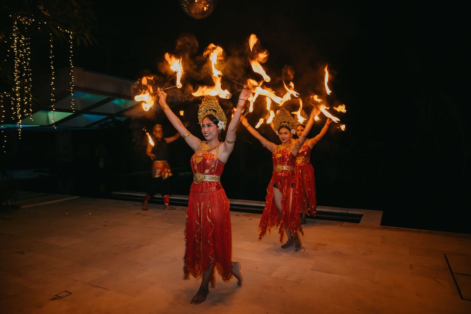 Fire dancers in red costumes perform with flaming props at an outdoor wedding reception at night.
