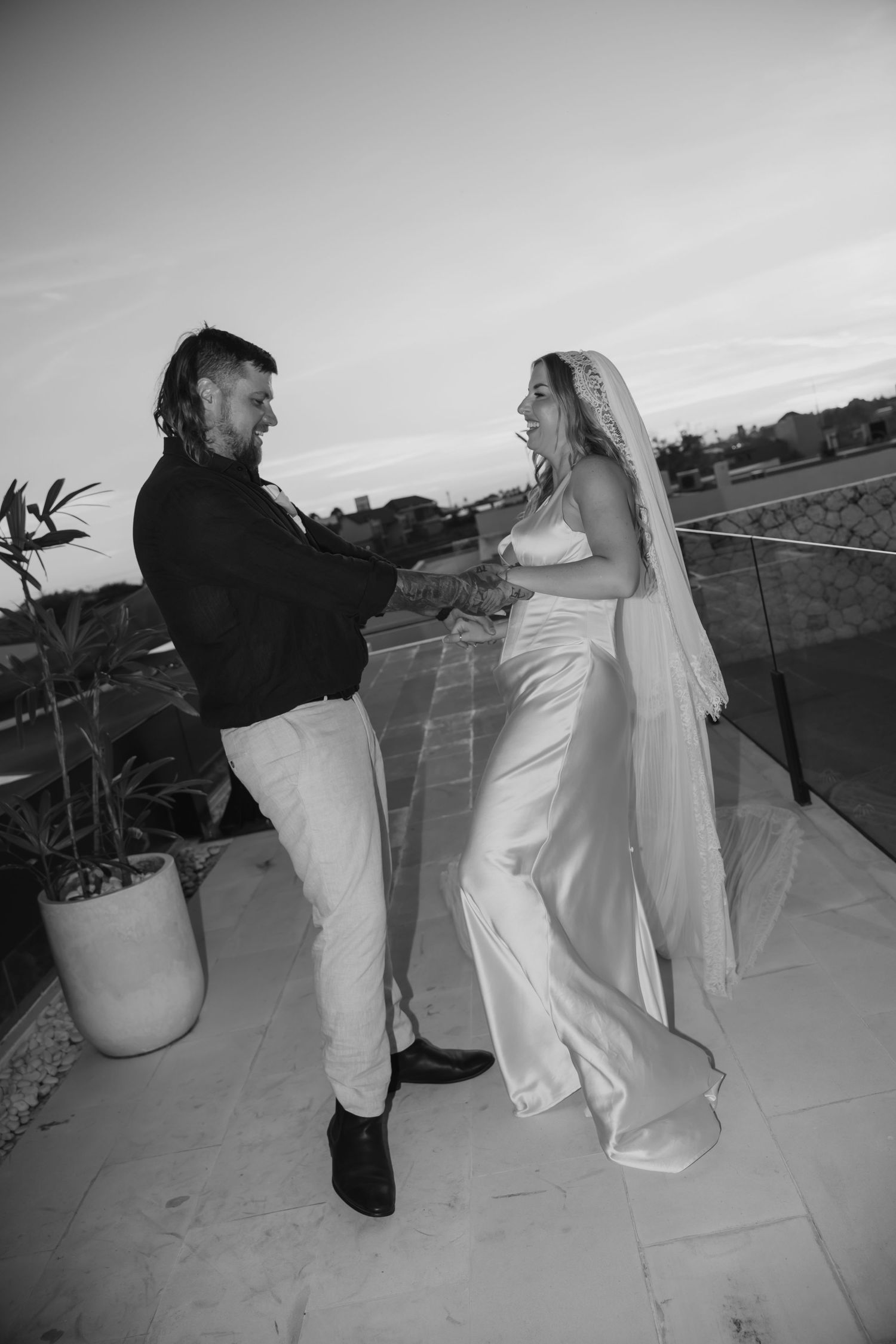 Black and white photo of bride and groom dancing joyfully on rooftop terrace at dusk.