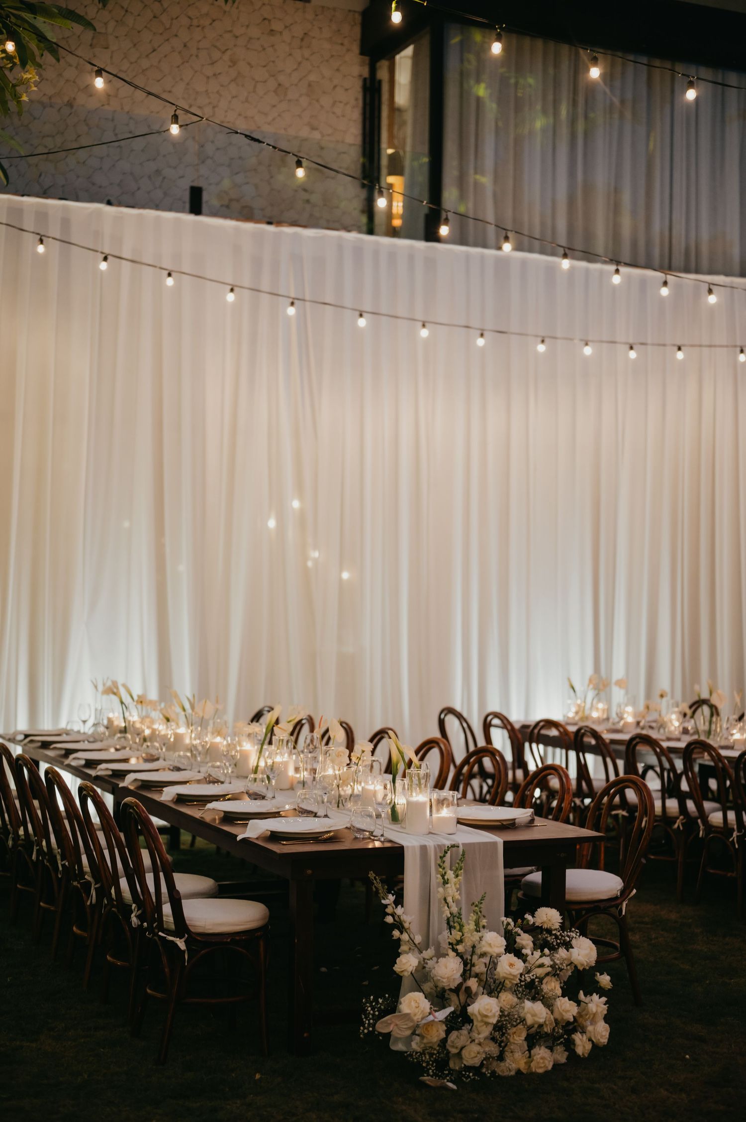 Elegant wedding reception with long wooden tables, white draping, string lights, and floral arrangements on grass floor.