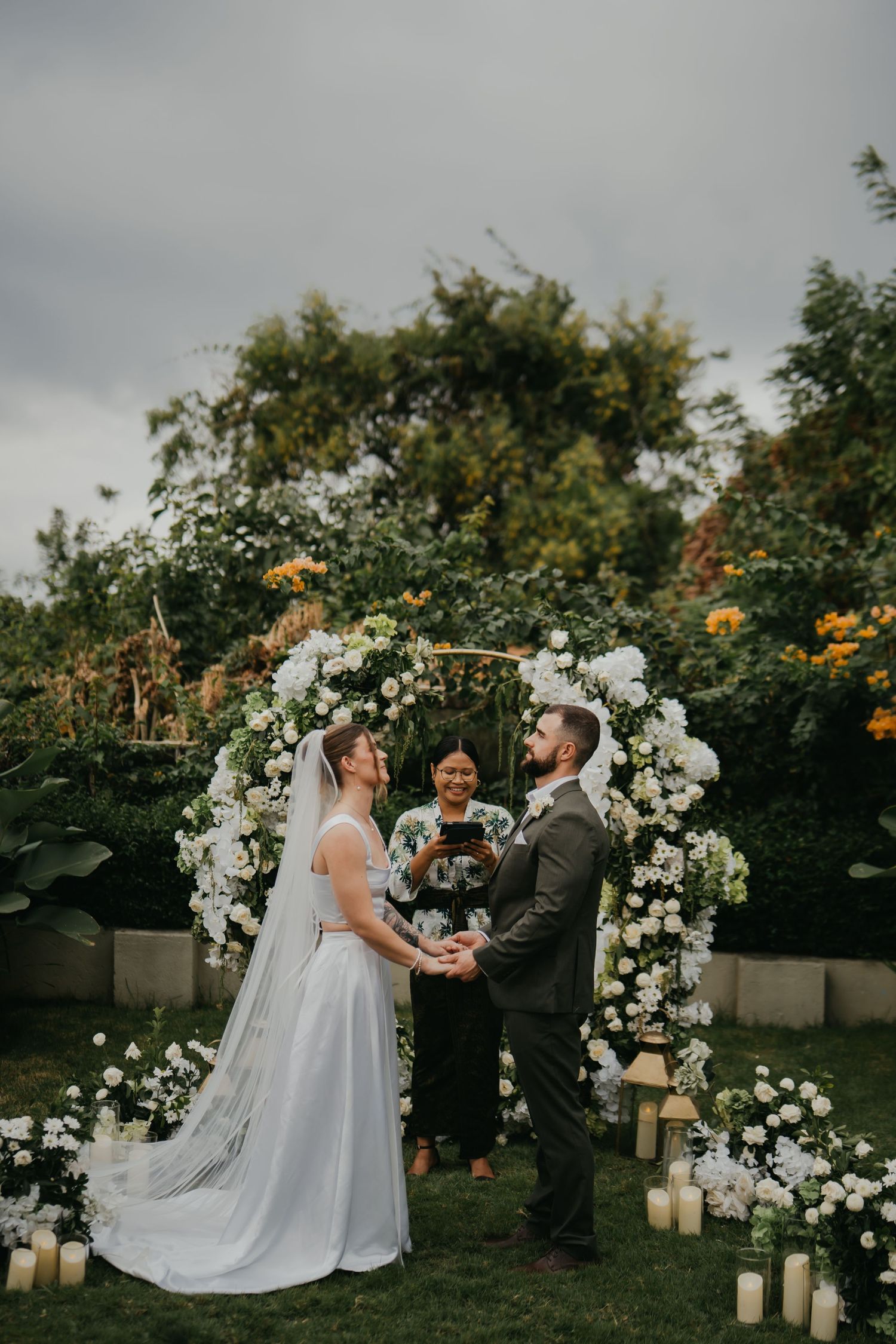 Couple exchanging vows at outdoor garden wedding ceremony beneath white floral arch with candles.