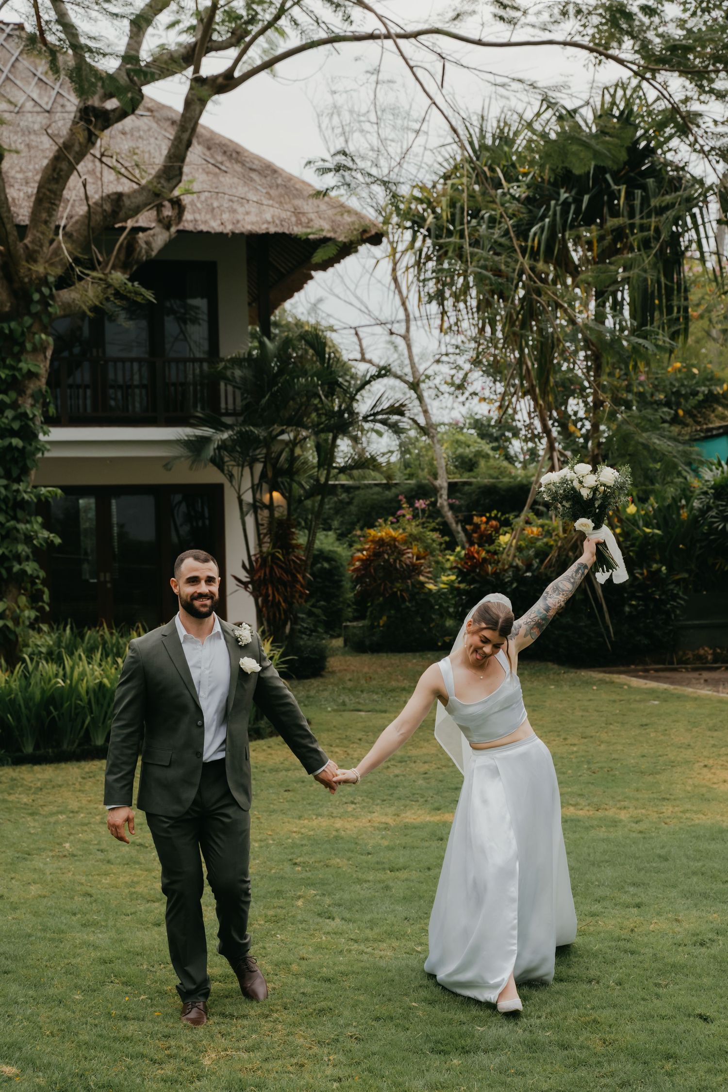 Joyful newlyweds hold hands walking through lush tropical garden venue.