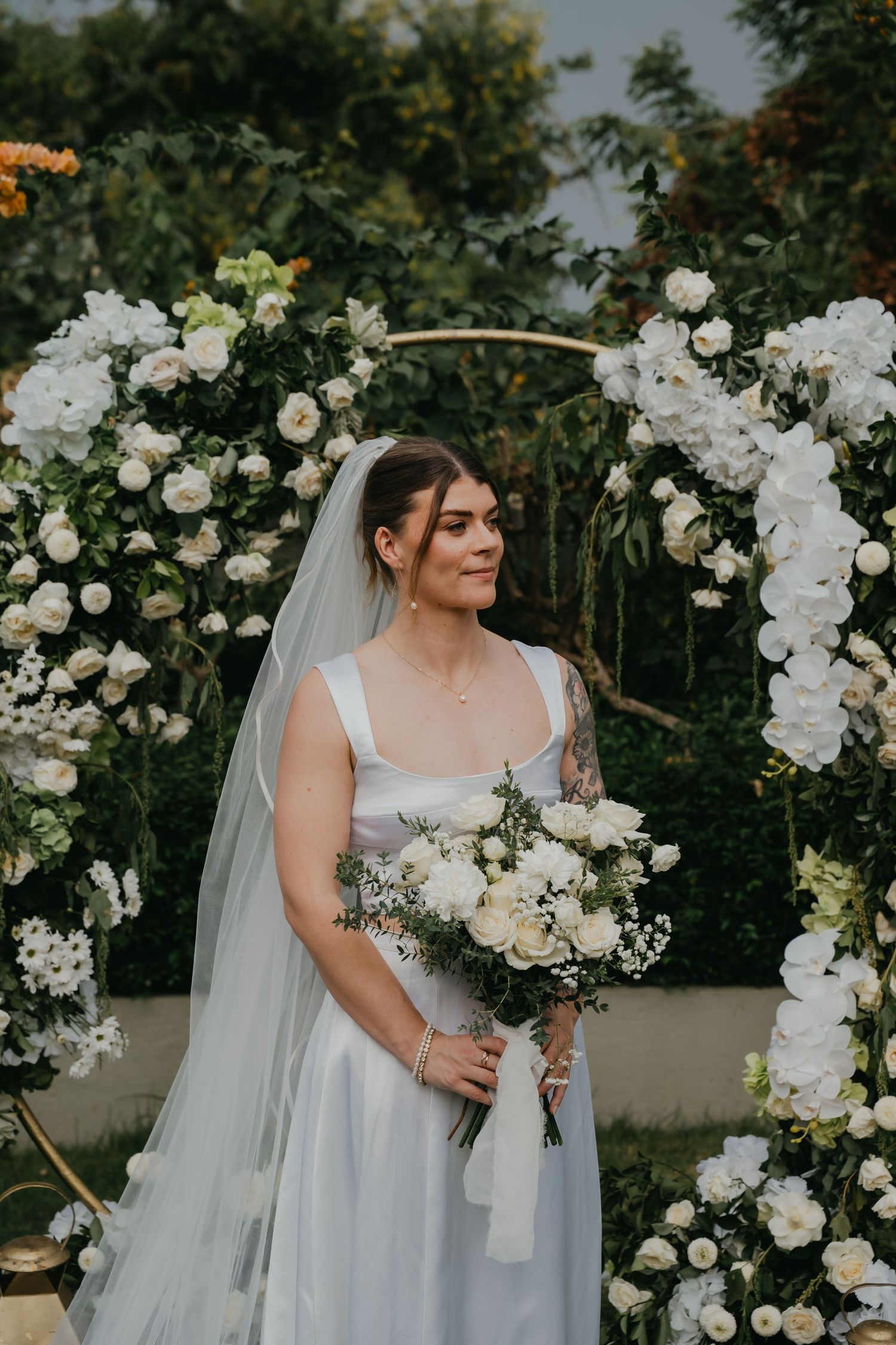 Bride in a white gown holds a bouquet of white roses, standing before a floral arch outdoors.