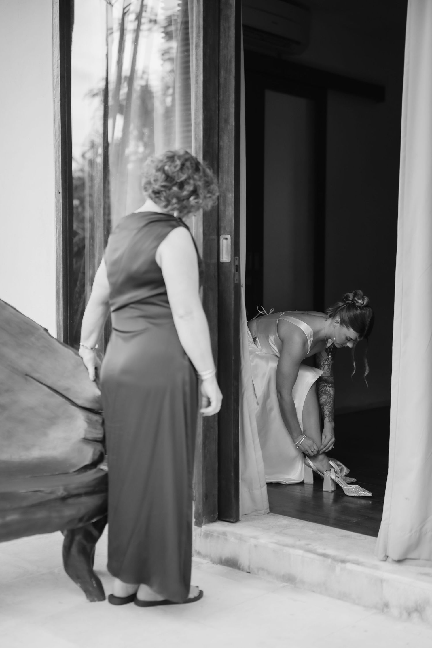 Black and white photo of bride putting on shoes in doorway while woman in elegant gown holds her dress train.