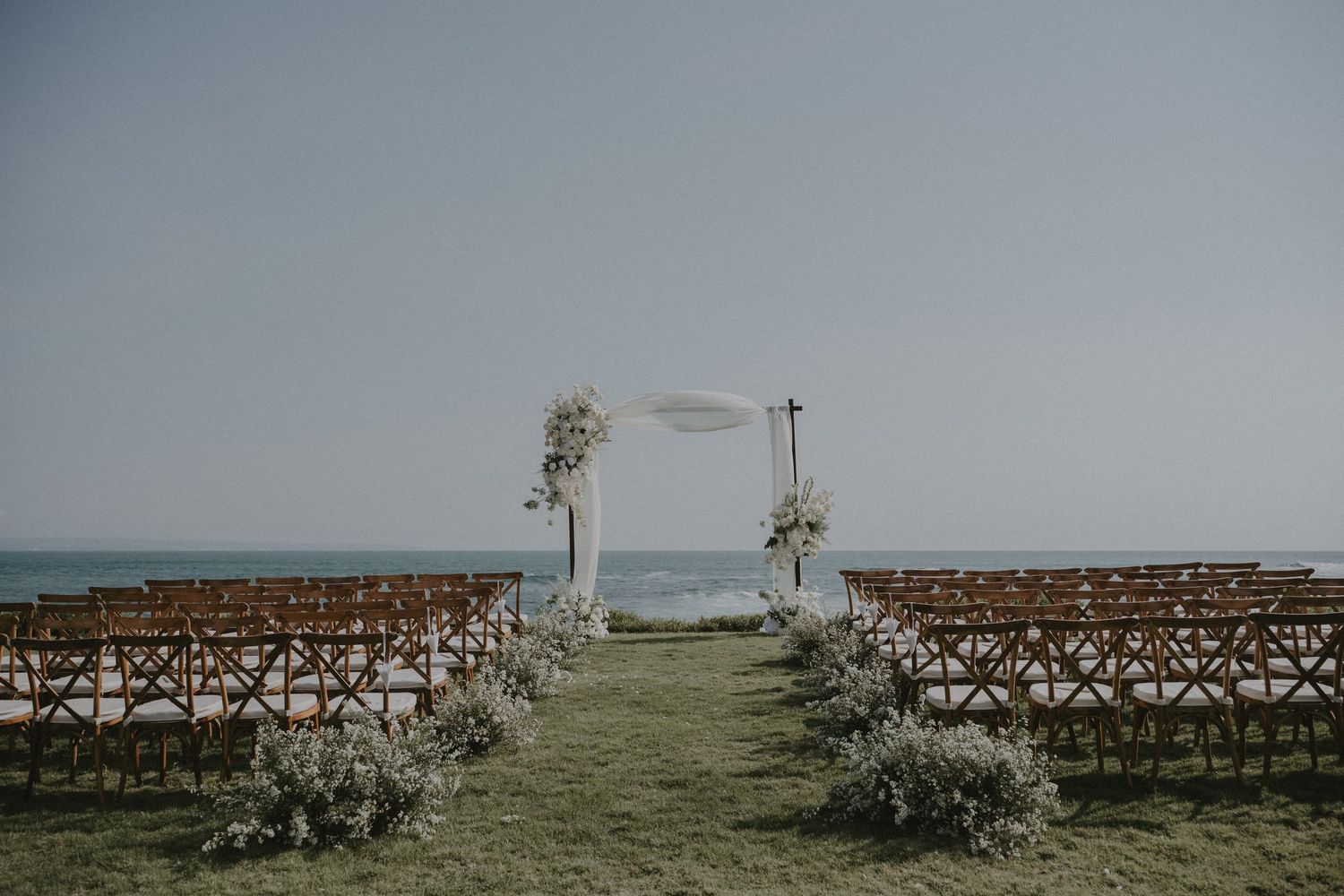 Rustic wooden chairs arranged in rows for an outdoor ceremony with ocean views and palm trees in the background.
