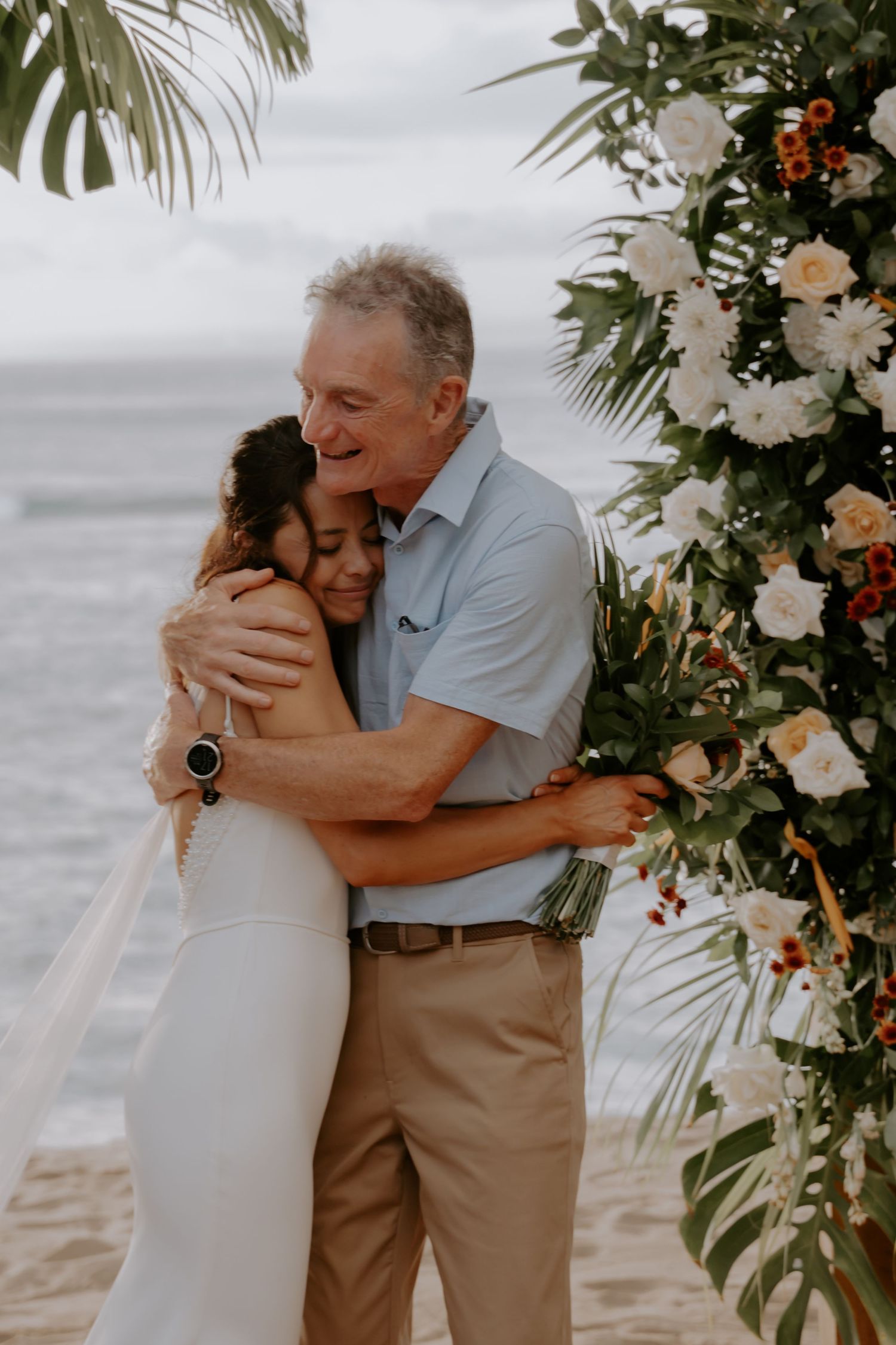 A tender embrace at a beach wedding ceremony with floral arch decorations and ocean backdrop.