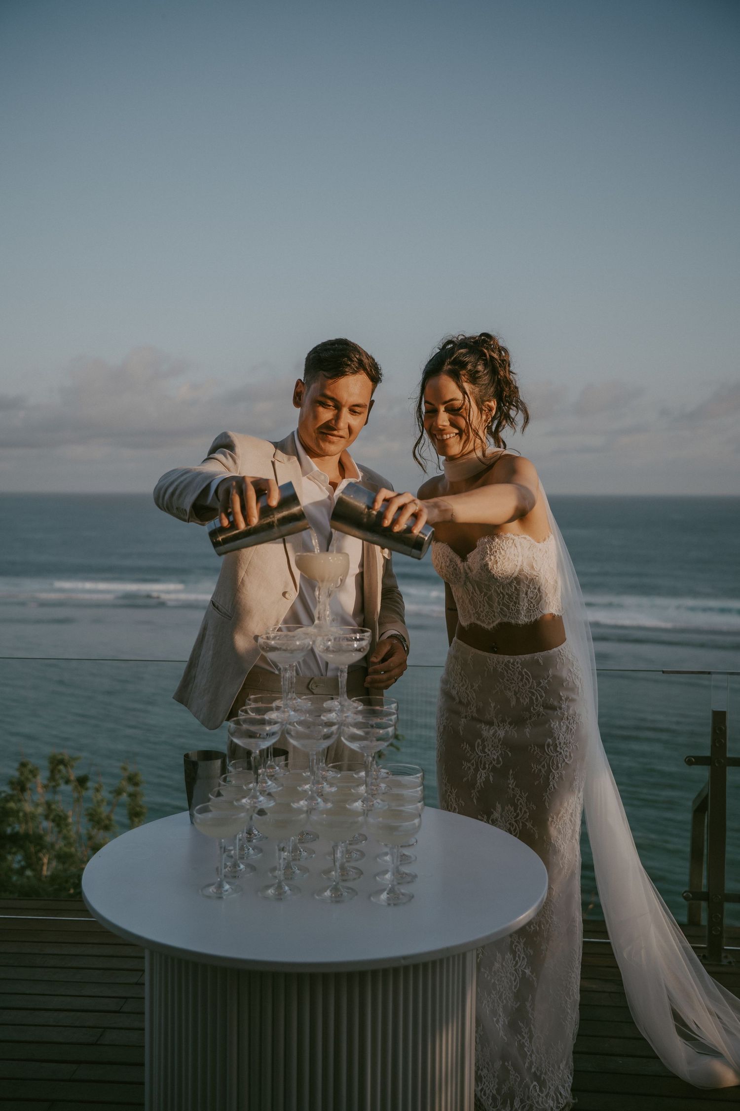 Two people in elegant attire pour champagne into a tower of glasses on a deck overlooking the ocean at sunset.