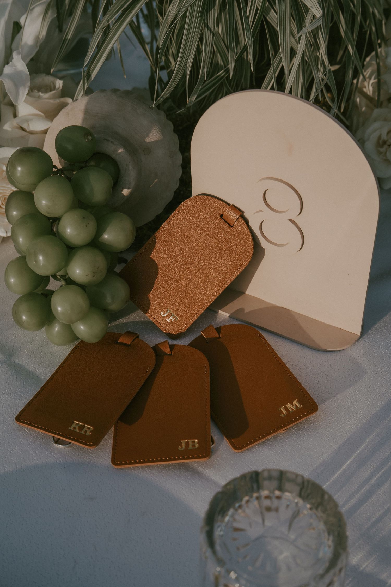 Brown leather travel accessories and luggage tags arranged with decorative grapes in a crystal vase on a light surface.