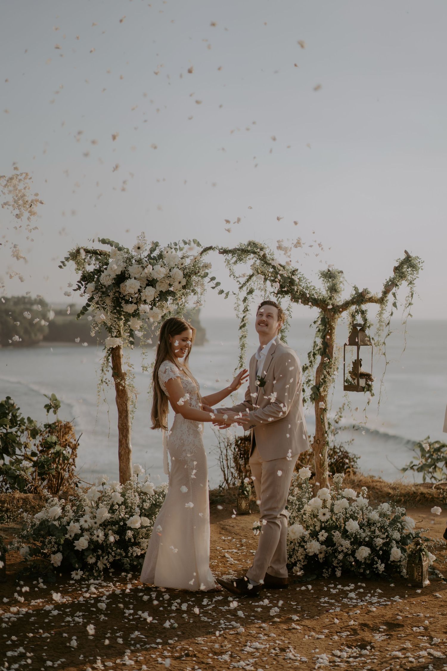 A romantic wedding ceremony takes place on a coastal cliff with flower petals floating in the breeze at sunset.