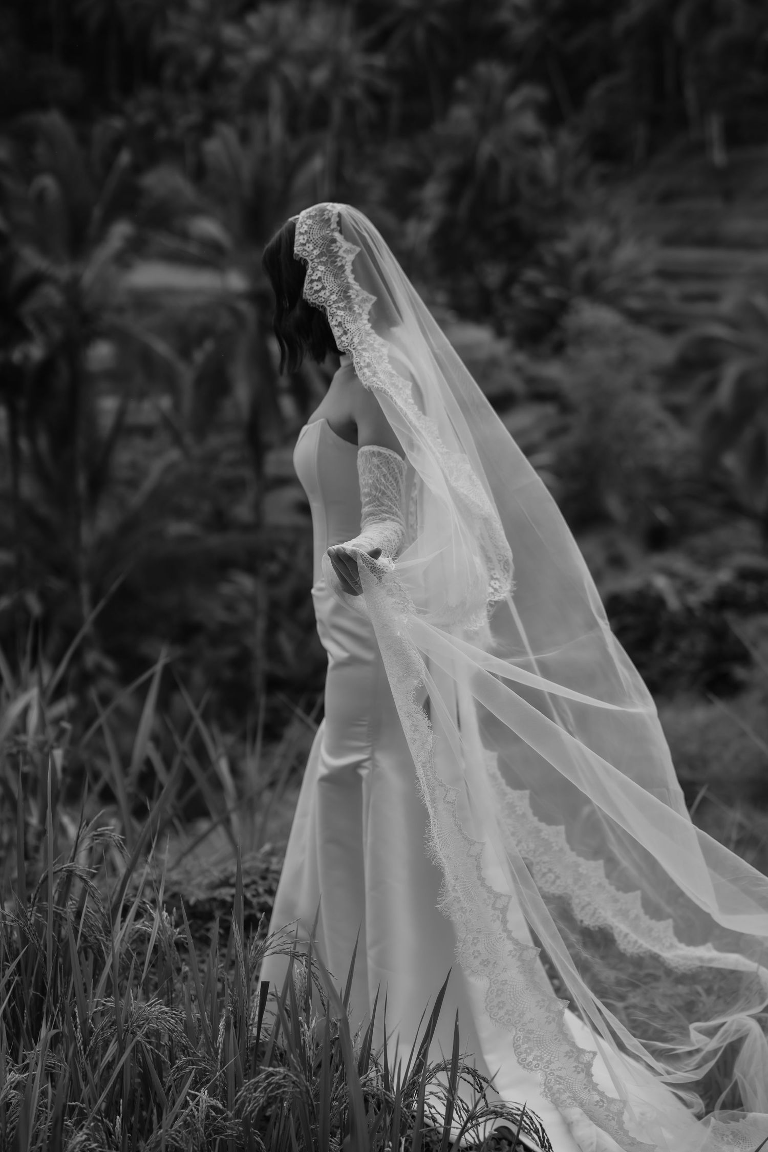 A flowing white wedding dress and veil move gracefully in the wind against a dark natural outdoor background.