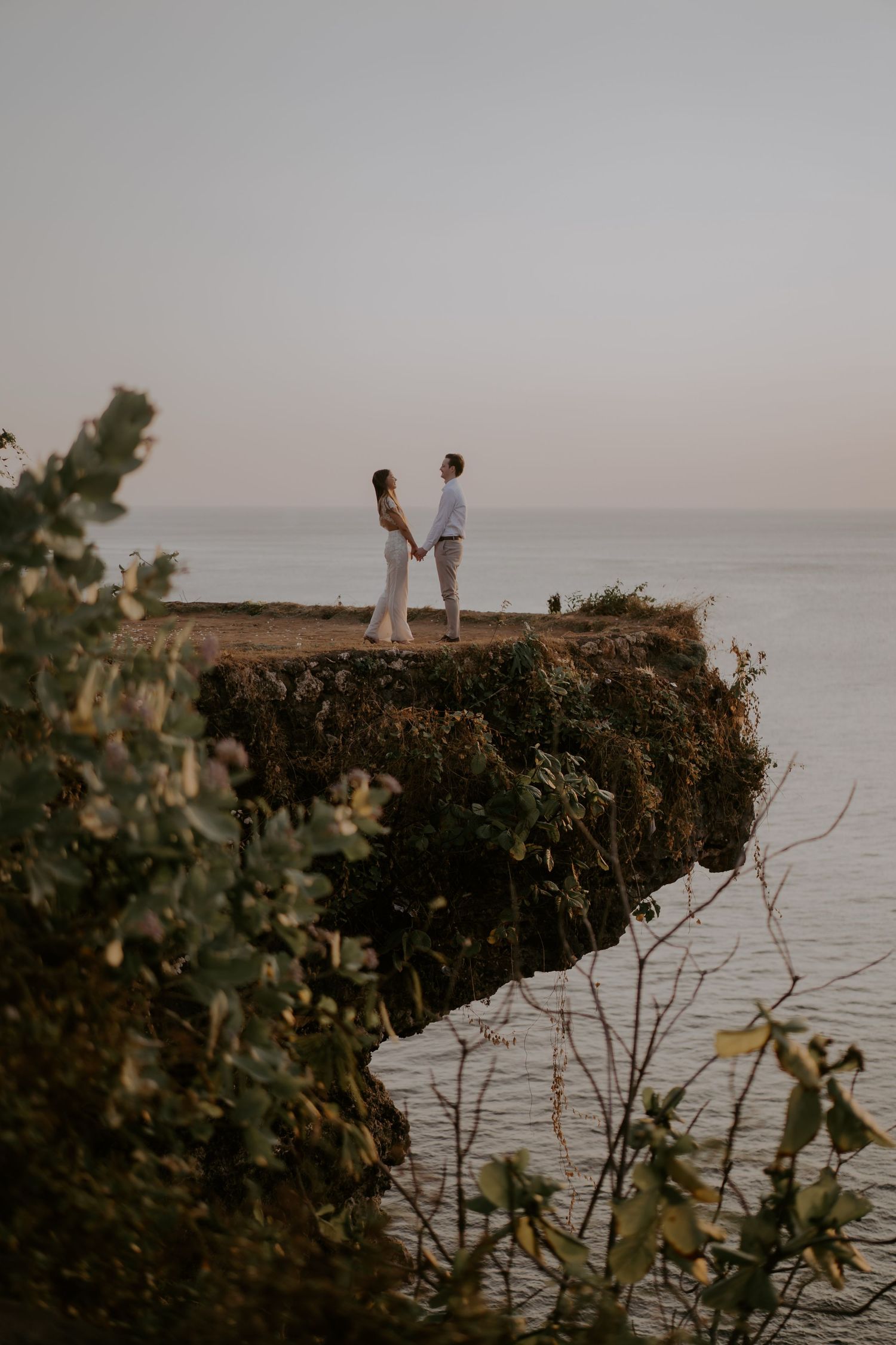 A couple stands on a rocky coastal cliff overlooking the ocean at sunset through a series of progressive shots.