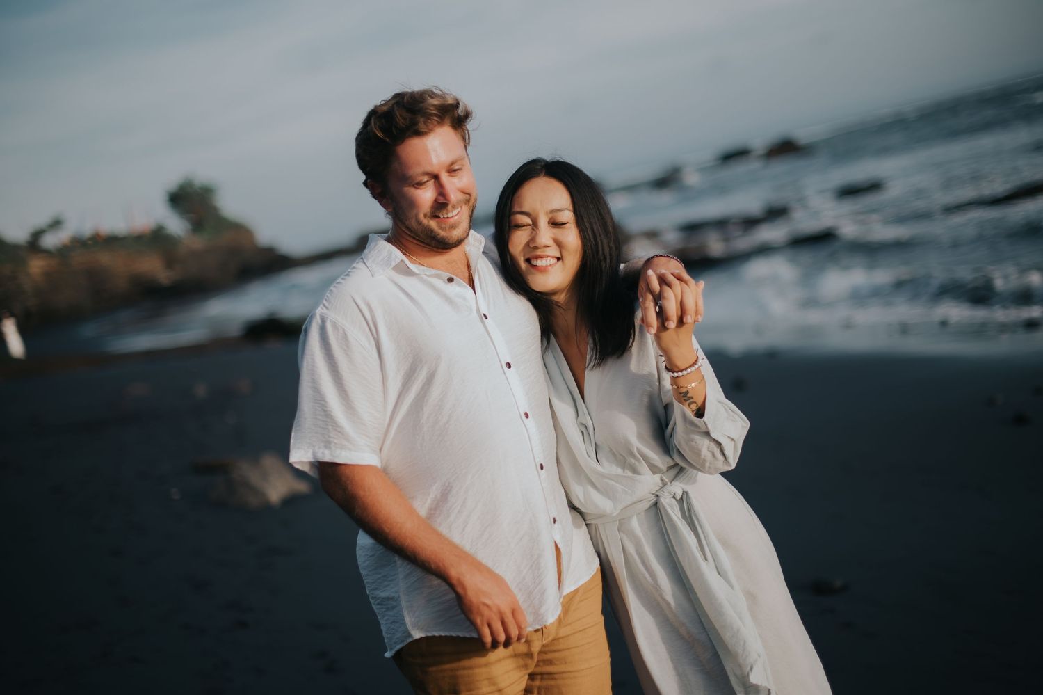 A couple shares a romantic moment together at sunset on a rocky beach with ocean waves in the background.