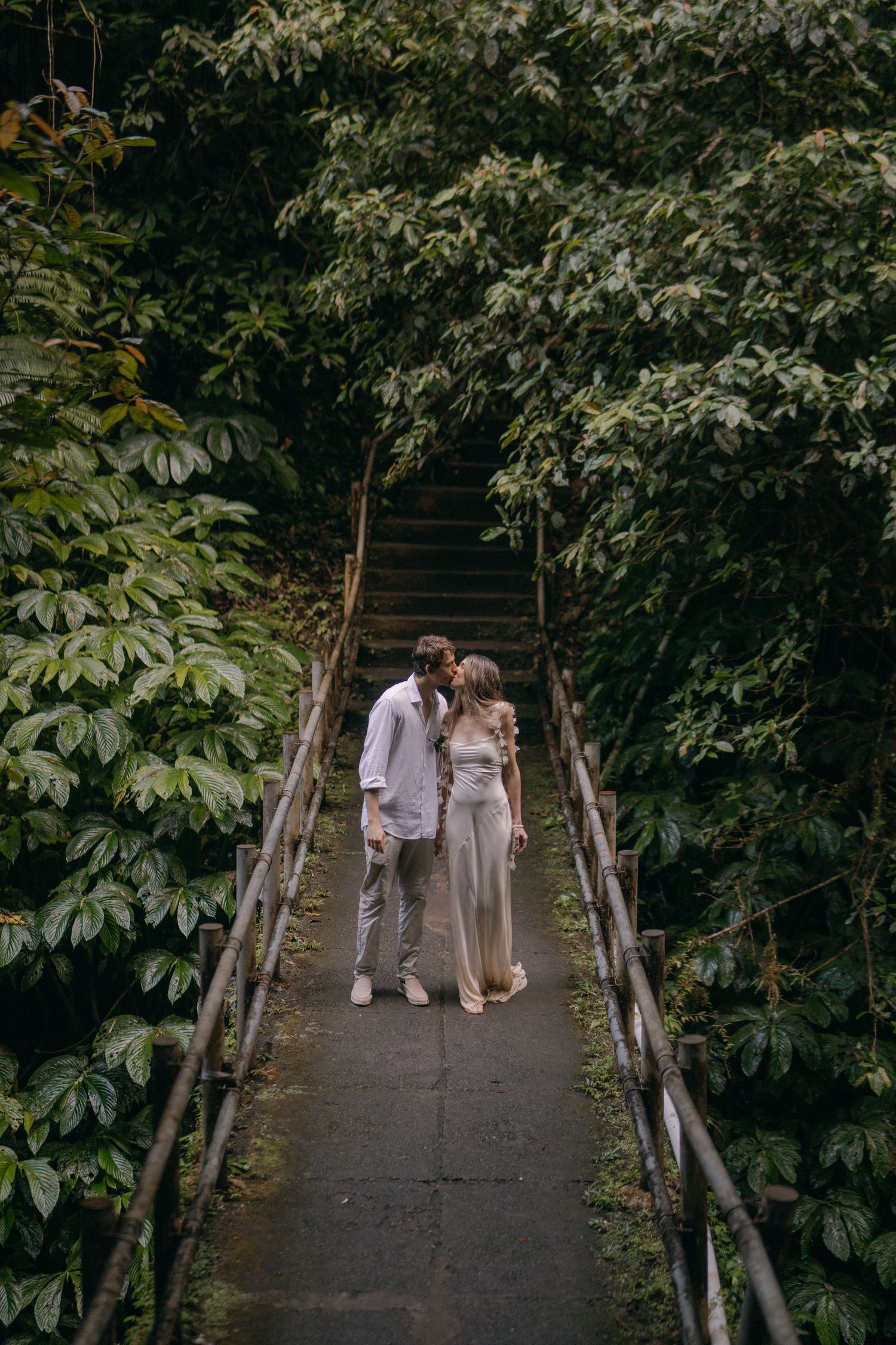 A couple in light-colored attire shares an intimate moment on a rustic wooden bridge surrounded by lush greenery.