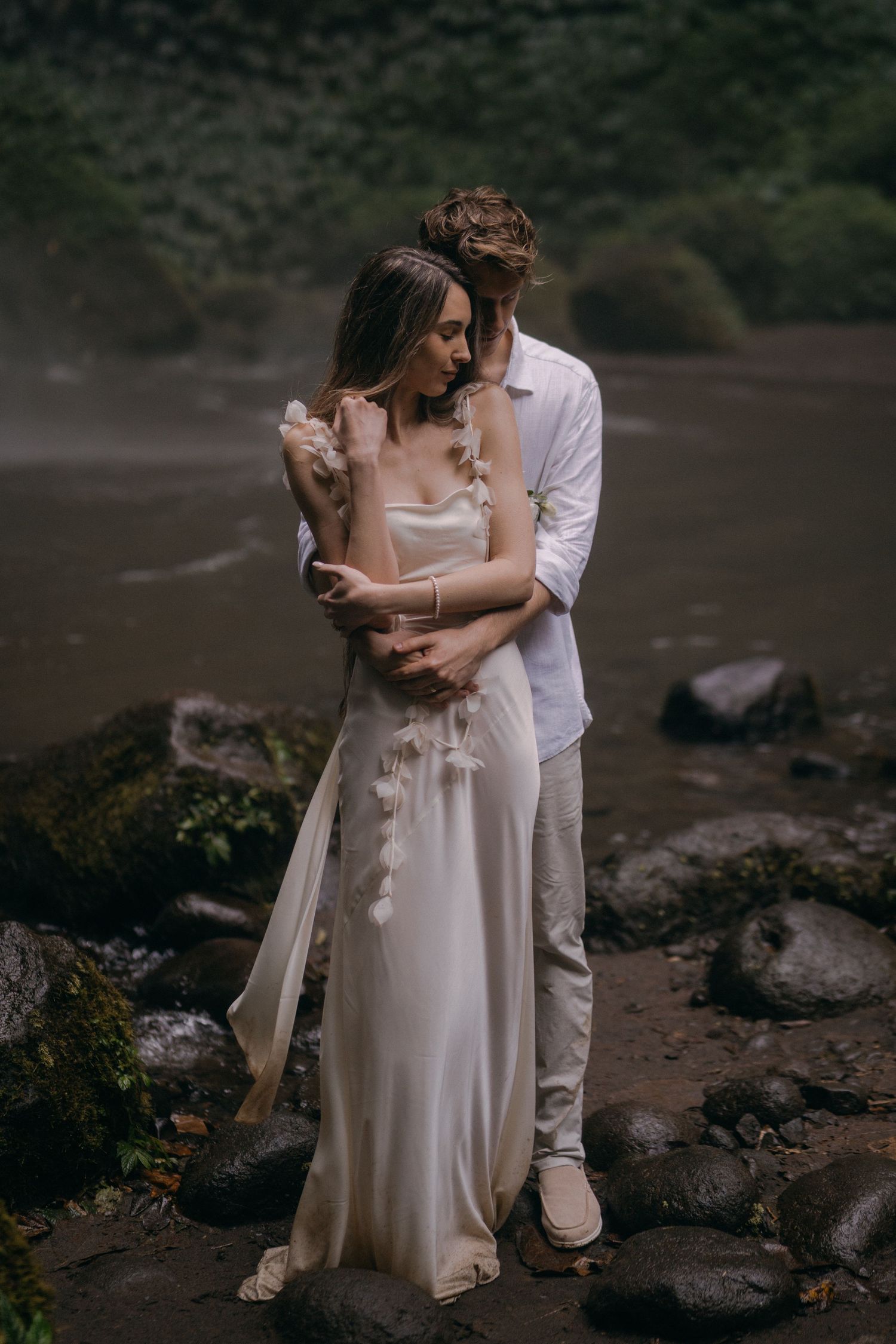 A romantic couple in white wedding attire embraces intimately on a rocky beach during a moody photoshoot.