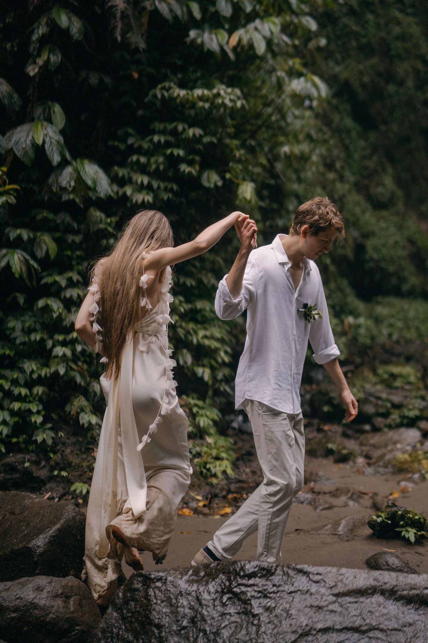 A couple in white outfits dances playfully on rocky terrain surrounded by lush greenery in a romantic photoshoot.