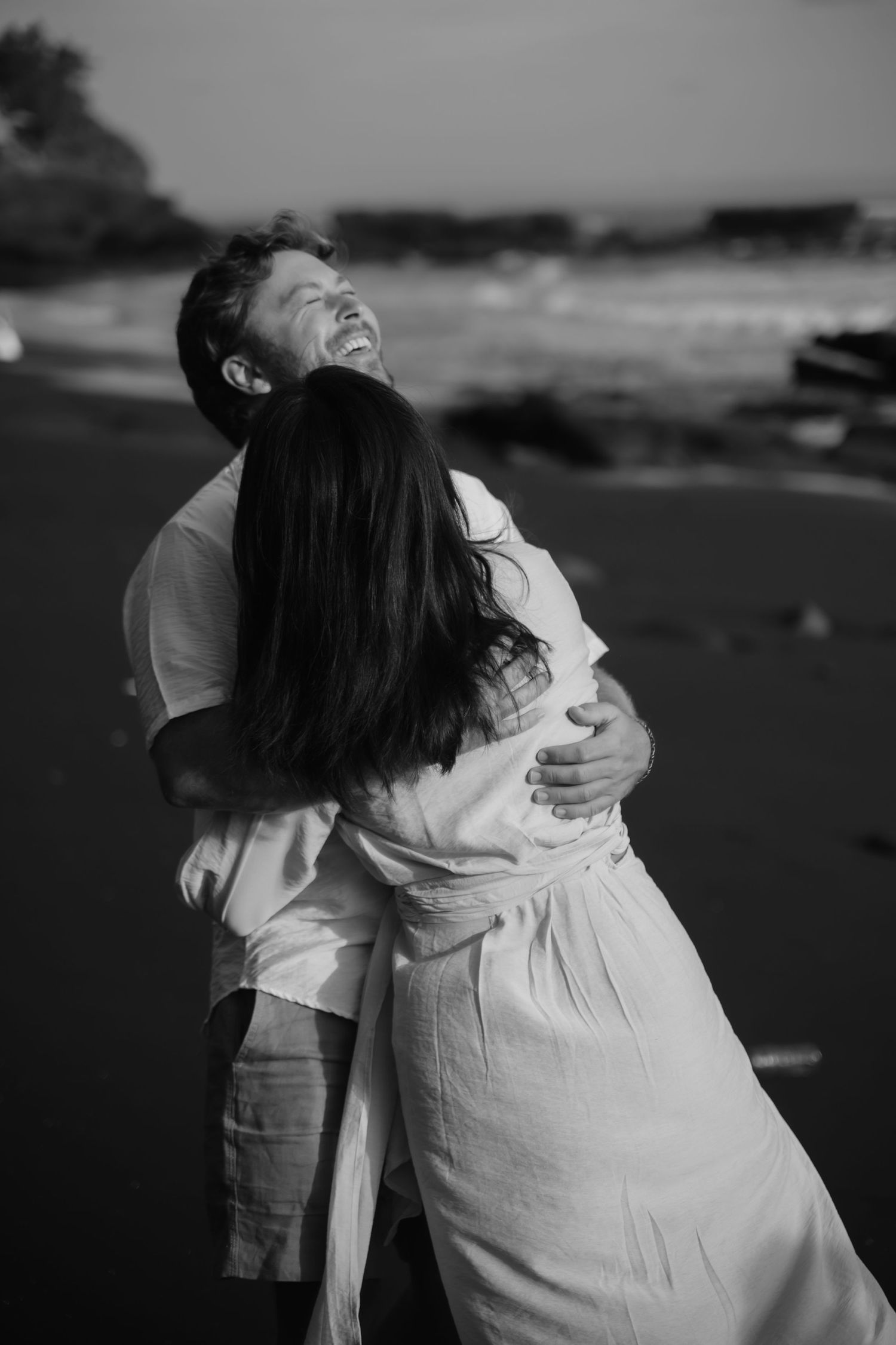 A couple in white clothing embraces and laughs together during a black and white photoshoot on the beach.